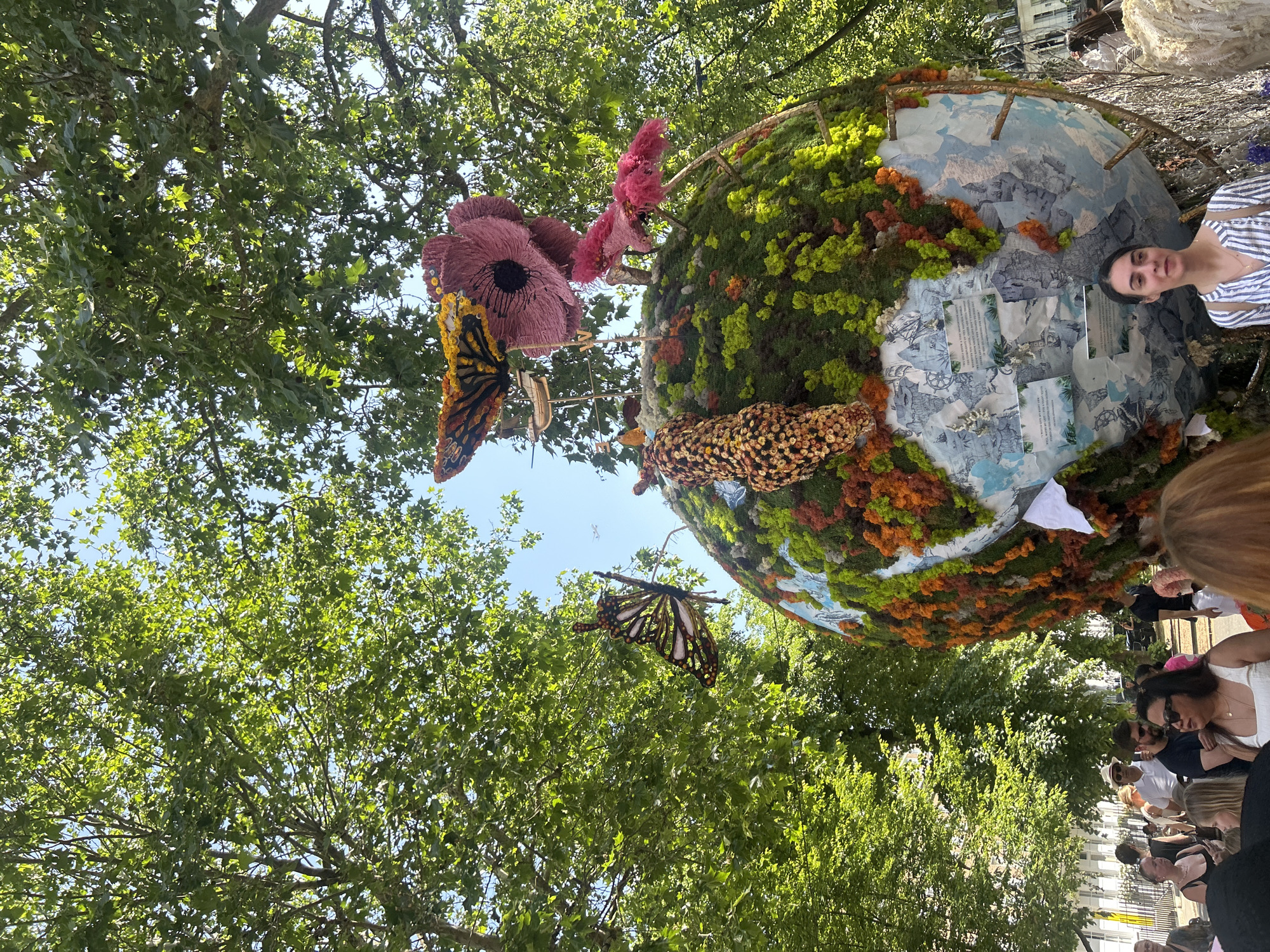 This image shows a public art installation in London, United Kingdom, featuring a large globe-shaped structure decorated with moss, flowers, and representations of landmasses. There are artificial butterflies attached to the structure, and the background includes a canopy of trees. Several people are visible in the foreground, suggesting that the artwork is in a public space.
