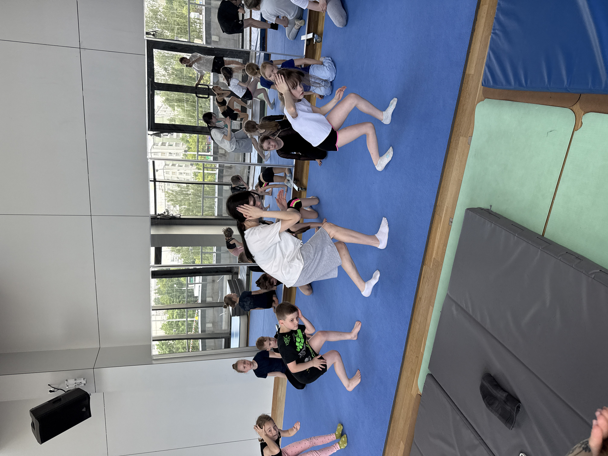 A group of children and adults are engaged in a physical activity session within an indoor facility, likely a gym or studio, located in Khimki, Russia. The setting is brightly lit by natural daylight, with large windows or mirrored walls reflecting the interior and revealing green trees and buildings outside, indicating a mild season.
Most participants, predominantly children, are seated on a large blue exercise mat, with many positioned in a manner suggesting core exercises or stretches, often with their hands placed behind their heads or on their hips. Their expressions appear focused on the activity. An adult in a white t-shirt and grey shorts stands towards the back, observing or instructing, while another adult in a black top is seated on the mat with the children.
The floor also features grey and light green exercise mats on the right, separated by a strip of wooden flooring. A black speaker is mounted on a white wall in the bottom-left corner of the image. On the t-shirt of a boy in the foreground, the text "X-SPORT" is partially visible. The scene depicts a structured group exercise or training session.
