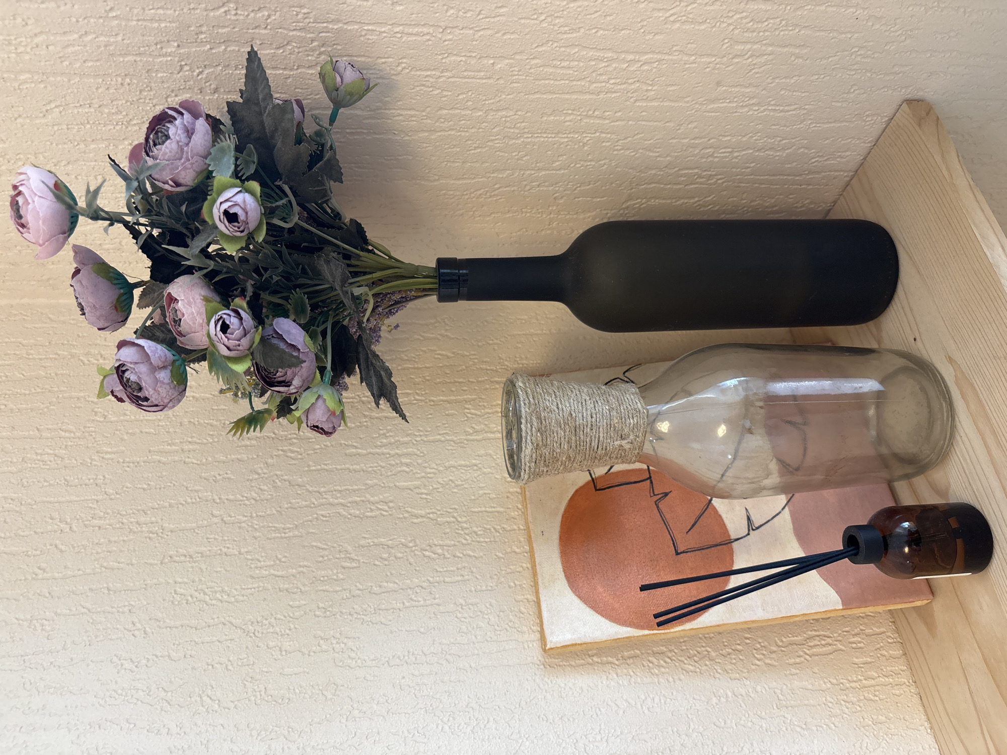 The image depicts a still life arrangement on a light-colored wooden shelf, against a textured, pale yellow wall. On the left is a small amber glass bottle with a black cap, containing dark liquid and three black diffuser reeds. To its right stands a clear glass bottle, its neck wrapped with twine. Behind these bottles, leaning against the wall, is a small canvas with an abstract design featuring a large orange circle and black lines. To the right of the glass bottle is a dark, matte black vase, holding a bouquet of artificial purple flowers with green leaves. The setting appears to be indoors, likely a home or decorative display area. There is no indication of Krasnodar, Russia, or any specific location within it.