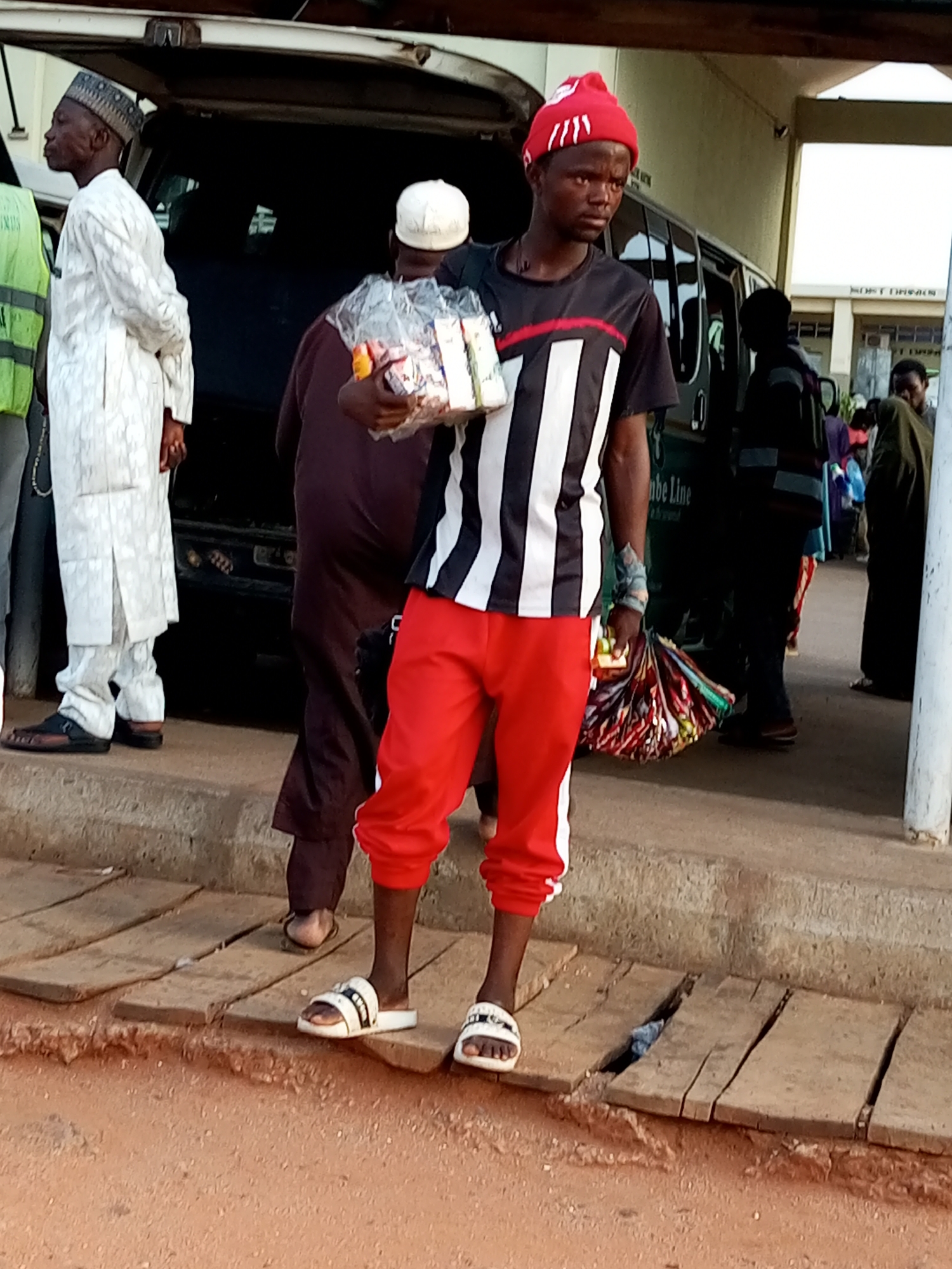The image shows a man standing on a wooden walkway in Gombe, Nigeria. He is wearing a red beanie, a black and white striped t-shirt, and red pants. He is holding a plastic bag filled with items and a bundle of colorful cloths. Behind him, a dark-colored van is parked. Other people, including a man in a white traditional outfit, are visible in the background. The scene appears to be outdoors, with a building in the distance. The walkway is made of weathered wooden planks laid over uneven ground.