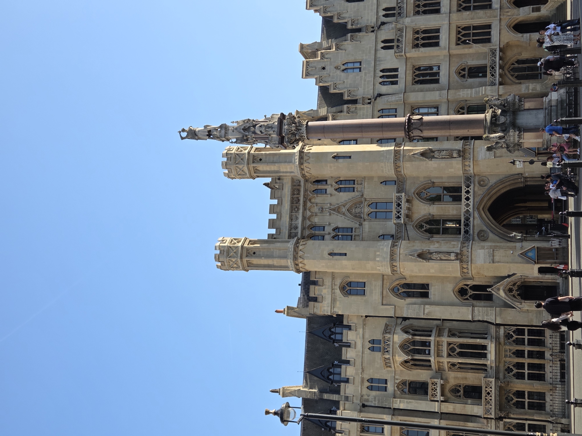 This image shows a view of the Victoria Tower Gardens, located in the City of London, United Kingdom. The architecture displays Gothic-style buildings. People are observed walking along the sidewalk. The sky is blue.