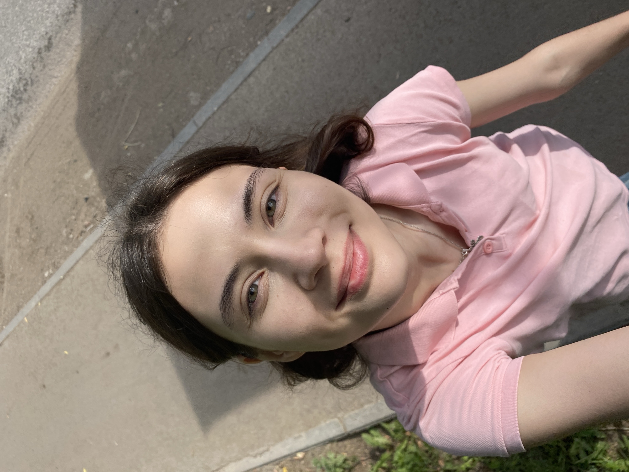 This is a selfie taken from a high angle outdoors. The subject is a young woman with long, dark brown hair and light skin. She is wearing a pink polo shirt and a silver necklace. Her eyes are green, and she is smiling. The background appears to be a sidewalk with some grass to the left and a shadow cast on the pavement. The lighting suggests it is daytime, likely sunny. The scene is consistent with a public area in Votkinsk, Russia, such as a park or walkway.