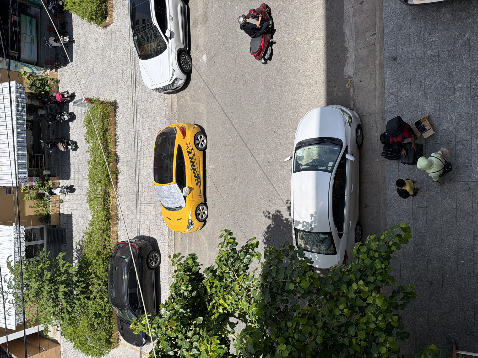 In the city of Bac Nha Trang Ward, Vietnam, on a sunny day, a street scene unfolds with various vehicles and pedestrians. A black SUV is parked on a tiled sidewalk adjacent to a building with awnings and greenery. Across the street, a yellow hatchback with "SPORT" graphics is parked, partially covered by a sunshade. Further down the street, a white SUV is parked. A person wearing a dark top and helmet is riding a red scooter. On the sidewalk, two individuals are standing near a white sedan. One person is wearing a yellow shirt and the other a light green hooded jacket. They are next to several pieces of luggage, including two suitcases and a cardboard box.