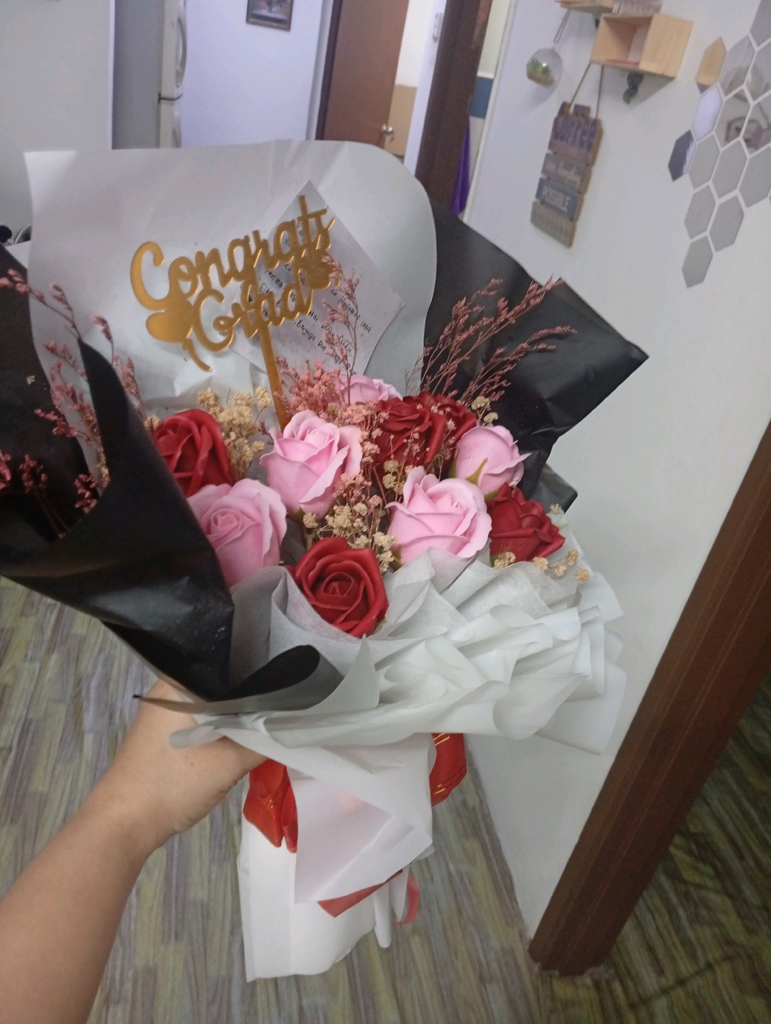 A close-up, slightly blurred, eye-level shot captures a bouquet of artificial roses, primarily pink and red, being held by a person's arm and hand. The bouquet is wrapped in black and white paper, with a gold-colored "Congrats Grad" cake topper prominently displayed. The background is out of focus, showing a wall with some decorative elements and a wooden floor. The scene appears to be indoors. This image is not in the city of Selayang Municipal Council, Malaysia.