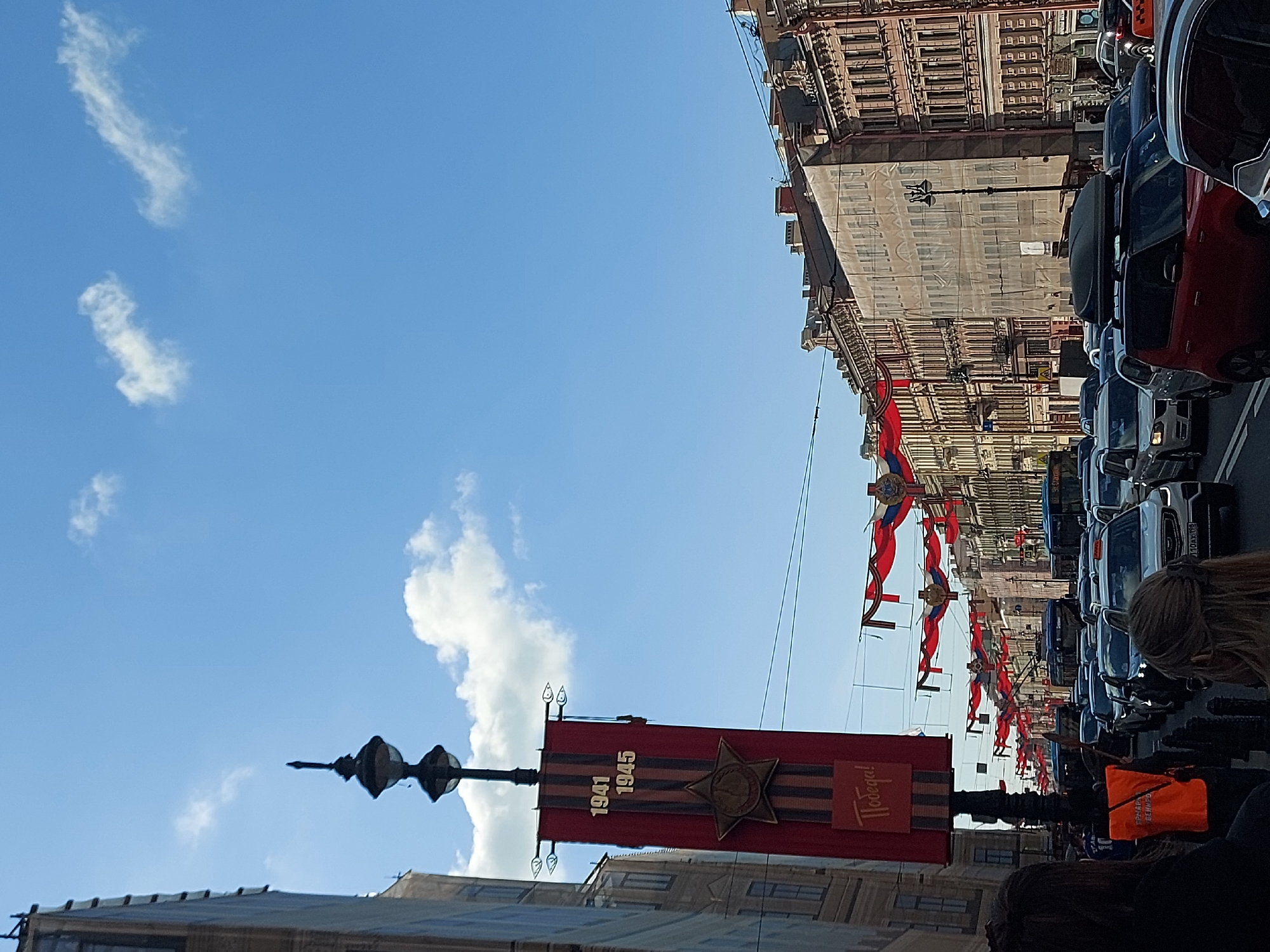 The image captures a street scene in Saint Petersburg, Russia, adorned with decorations for Victory Day. A red banner with the inscription "1941 1945" and a star emblem hangs from a lamppost. Above, red and white flags are strung across the street. The background features buildings and a line of cars, indicative of a street. Several people are visible in the foreground. The sky is clear blue with some clouds.