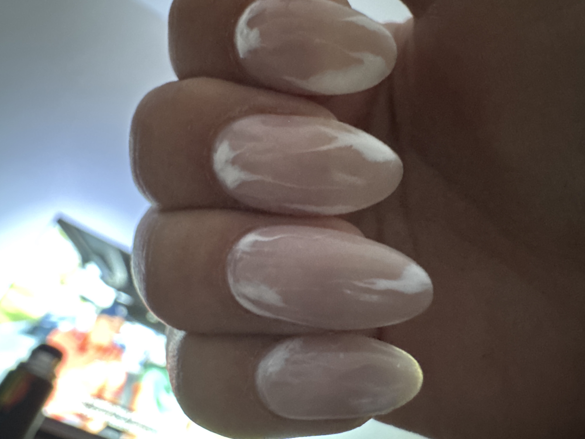 The image shows a close-up view of a person's hand with light pink almond-shaped fingernails that have a marbled white design. The hand is positioned vertically in the frame, with the fingers slightly curled. The background is out of focus, but appears to be a brightly lit area with some colorful elements, possibly a digital screen or artwork. The lighting suggests an indoor environment. There is no discernible indication of the location being San Fernando del Valle de Catamarca, Argentina.