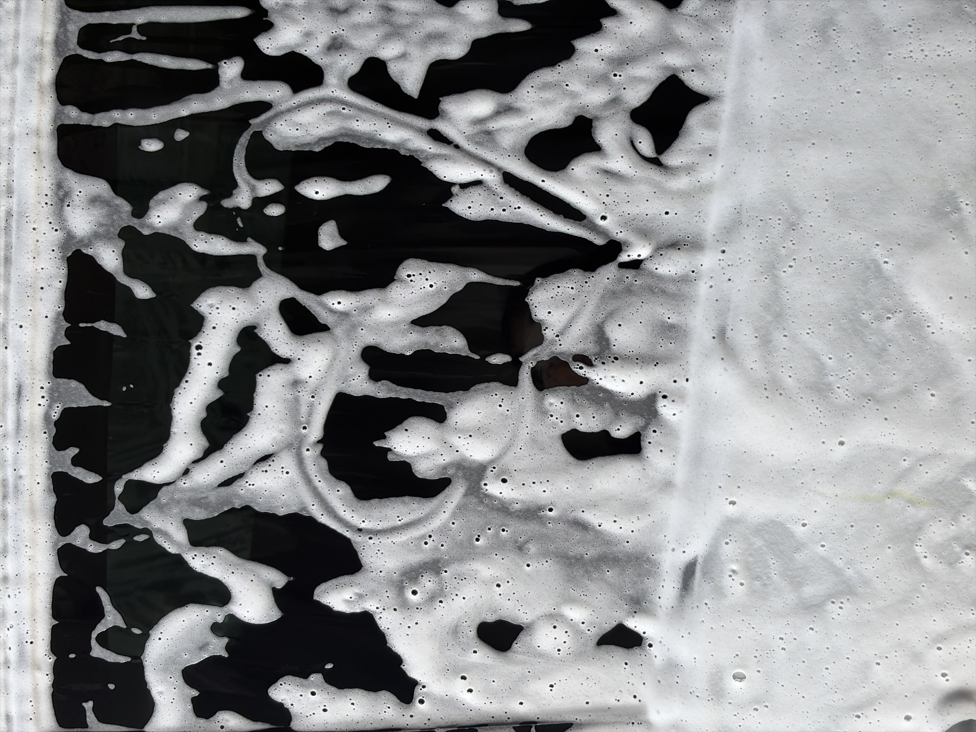 The image shows a glossy black surface partially covered with white foam. The foam has a bubbly texture with varying degrees of thickness, transitioning from dense coverage on the right side to scattered patches on the left. The black surface is visible through the foam, creating a contrasting pattern of light and dark areas. The location is unknown.
