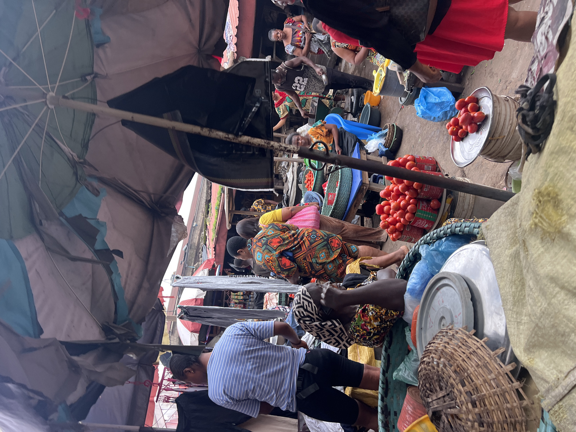 This is a medium shot, eye-level, outdoor photograph taken at the Nkpor market in Nigeria. The scene is crowded, with several people visible amidst stalls and awnings. A large, partially collapsed umbrella dominates the foreground and midground, casting shadows and obscuring parts of the scene.
In the midground, a woman wearing a headscarf and a colorful patterned dress is seated, speaking on a mobile phone. To her left, a person wearing a blue and white striped t-shirt and black shorts stands looking away from the camera. Behind the seated woman, several other individuals are visible, including a woman in a vibrant, geometrically patterned top.
The market appears to be selling produce, with baskets and crates of tomatoes prominently displayed. Several metal bowls and buckets are also present. The ground is dirt and the stalls are constructed from wood and tarpaulin. The overall atmosphere suggests a busy, active market environment.