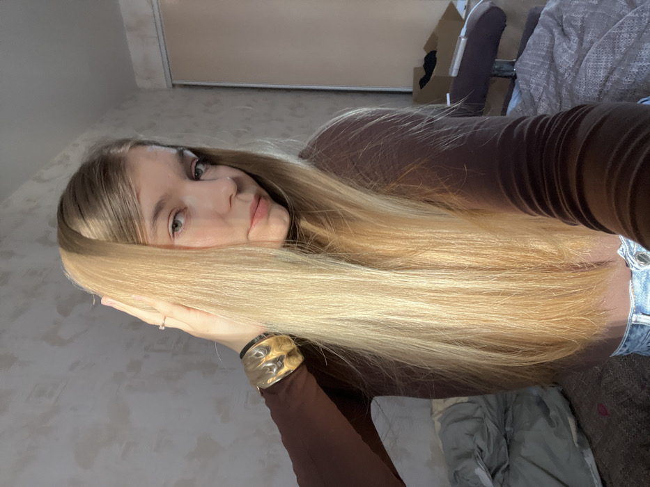 A blonde woman with long, straight hair poses in a selfie. She wears a brown long-sleeved shirt. She is wearing a large, gold cuff bracelet and a ring. Behind her is a bedroom with a wardrobe and a couch. The photo is rotated at an angle.