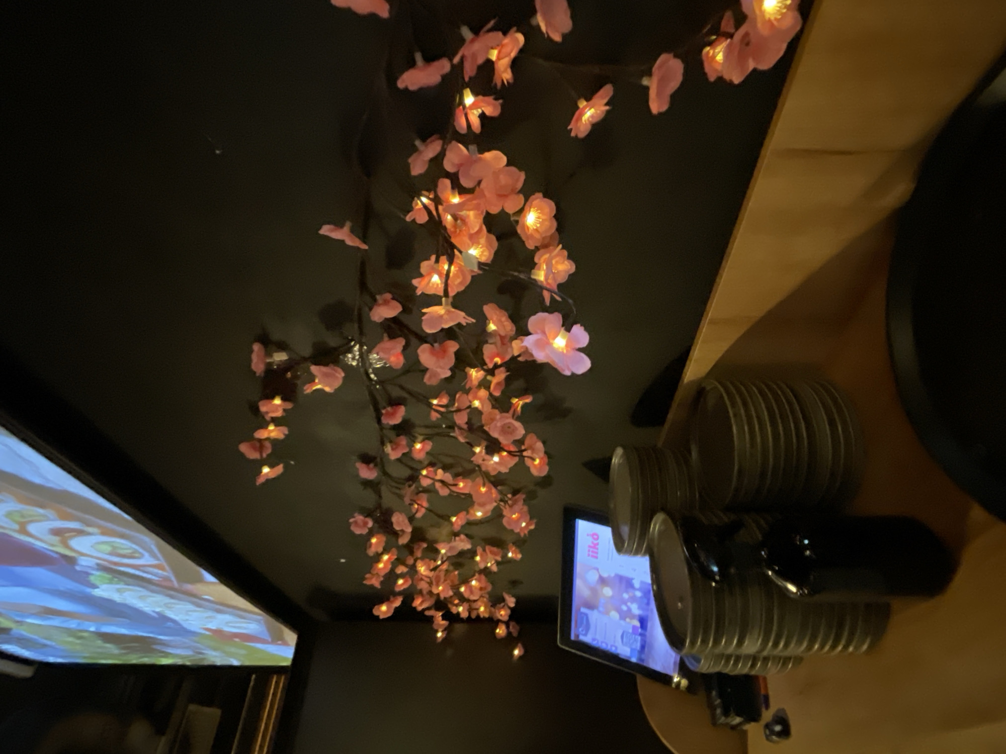 The image depicts a dimly lit interior, possibly a restaurant or bar, with a dark wall as the primary backdrop. A string of artificial cherry blossoms with integrated warm lights is draped across the wall, creating a decorative effect. In the foreground, on a wooden surface, are several stacked metallic containers, a tablet displaying a colorful menu or advertisement with the text "iikó" visible, and a dark spray bottle. Above the scene, a large projection screen displays a blurred, colorful image, suggesting a visual display is active. The overall ambiance is cozy and potentially atmospheric, enhanced by the soft glow of the decorative lights and the screen. The scene is devoid of any visible people or explicit actions. The location is identified as Moscow, Russia.