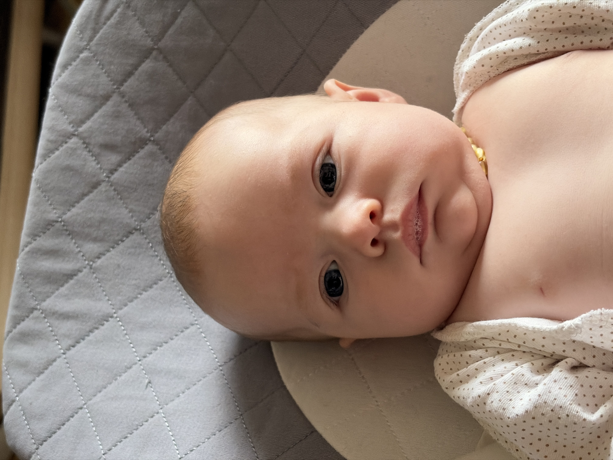 A baby lies on a padded gray surface, possibly a quilted mat or cushion. The baby is fair-skinned with dark eyes. They are wearing a light-colored top with small dots and a yellow beaded necklace. The background is out of focus. The location is not discernible from the image.
