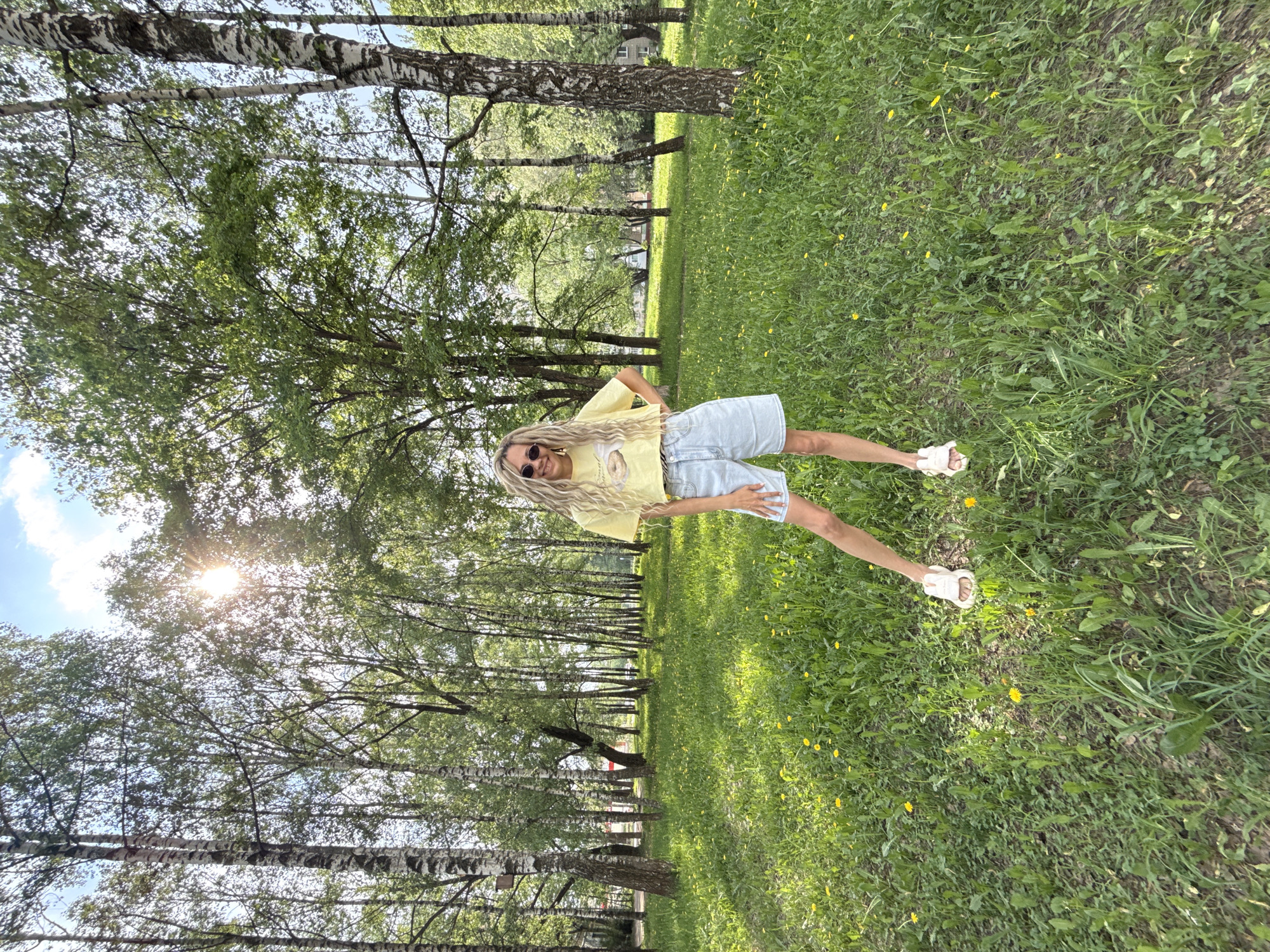 A full-length photograph shows a woman standing in a grassy field with trees in the background. The woman has long blonde hair and is wearing sunglasses, a yellow t-shirt, denim shorts, and white sandals. She is smiling and has her hands on her hips. The field is green with yellow flowers. The trees are birch trees with white bark. The sun is shining through the trees. The location is Rusanovo, Russia, with the typical birch tree coverage in a park.
