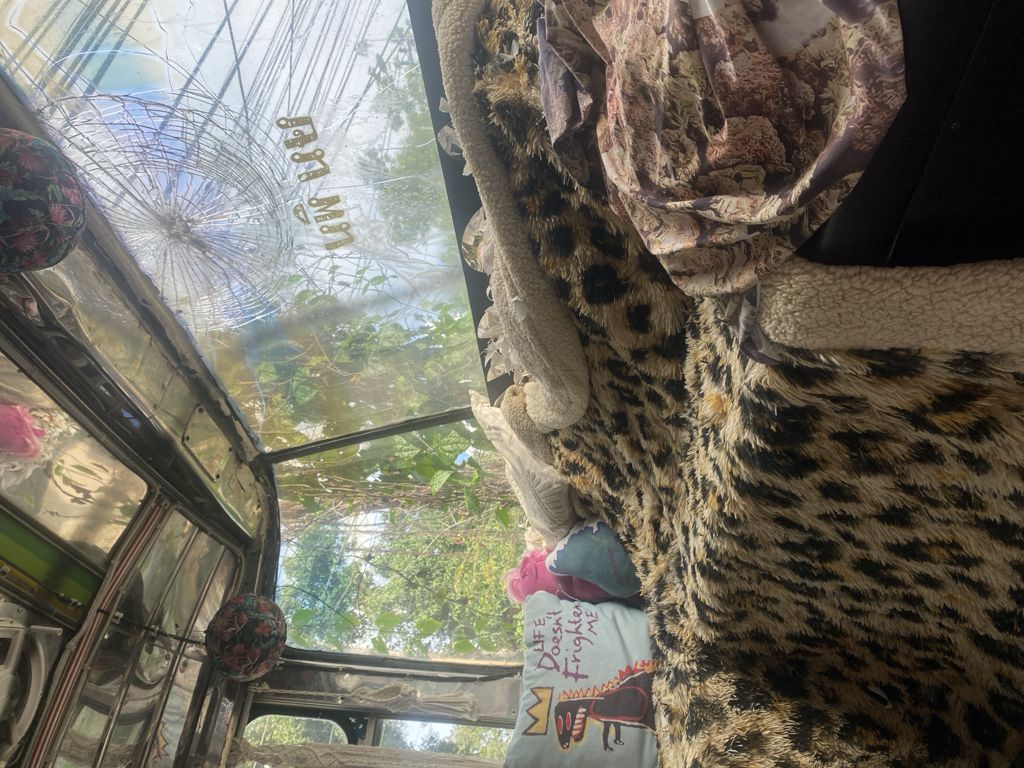 The image shows the interior of a vehicle, likely a tuk-tuk, in Thong Sala, Thailand. The windshield is cracked with a spiderweb pattern and Thai script visible on the glass. The interior is heavily decorated with various textiles, including a leopard print fabric and a pillow with an artistic drawing. Green foliage is visible outside the windows.