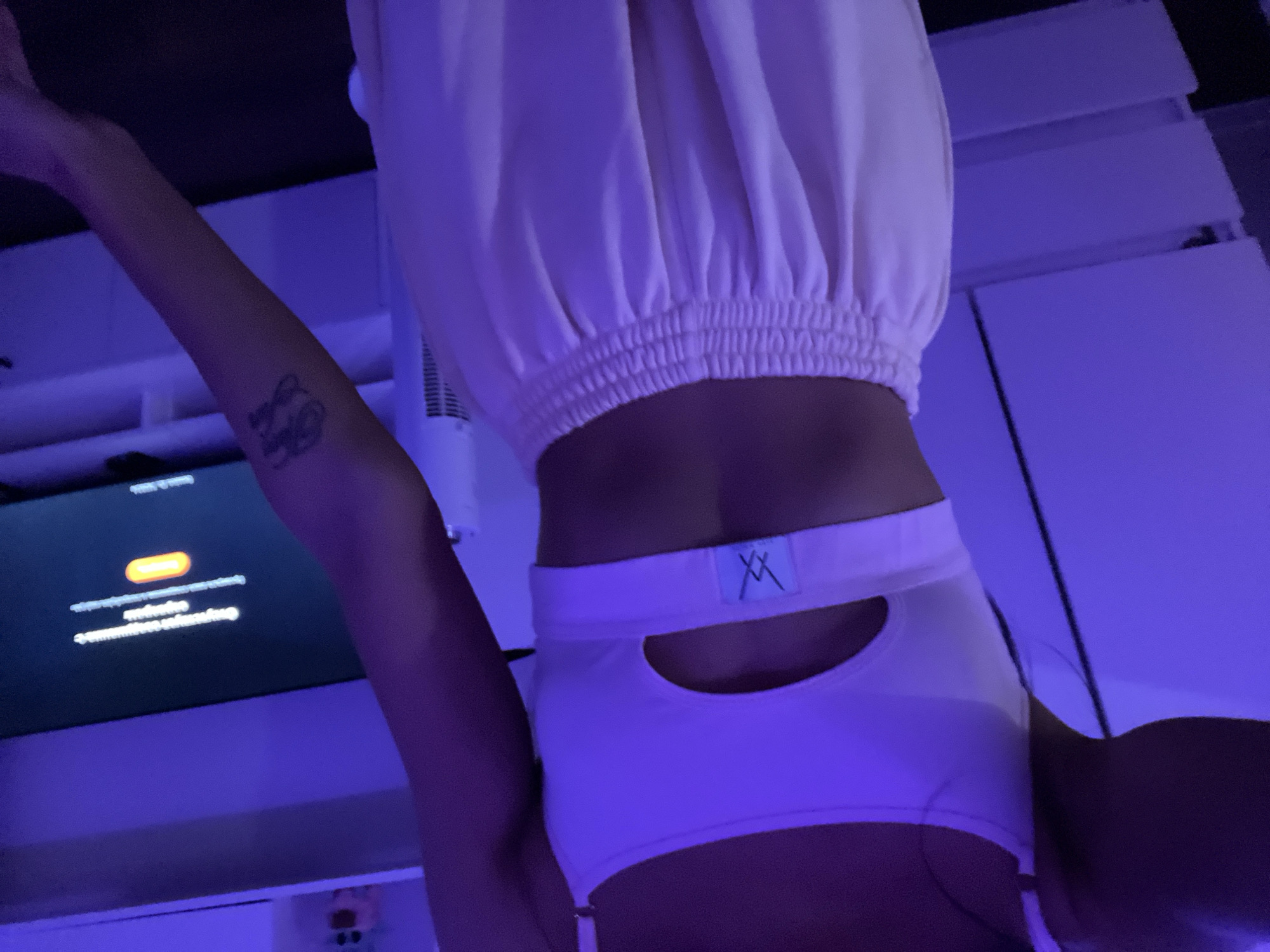 A person, seen from the chest down, is standing in a room illuminated by purple lighting. They are wearing a light-colored sports bra with a cut-out detail in the center and light-colored sweatpants with an elastic waistband. On the person's left arm, there is a tattoo. In the background, a television screen displays an orange button with text in an unknown language. Other objects in the room include what appears to be shelving and a white fan. The location is unknown.