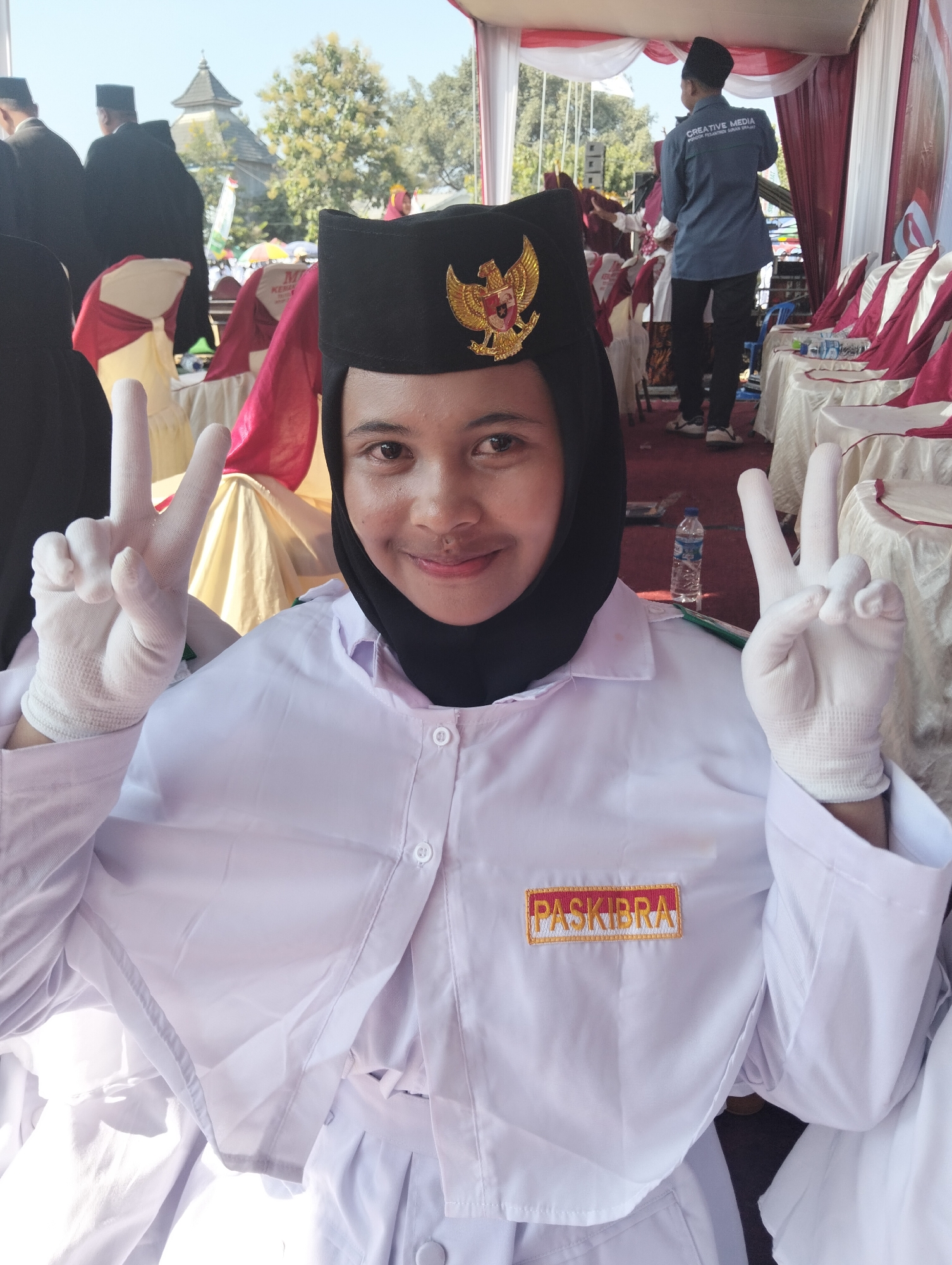 A young woman, wearing a black PASIKBRA hat adorned with the Indonesian coat of arms and a white uniform with white gloves, makes a peace sign with her right hand. She is positioned in the foreground, looking directly at the camera with a slight smile. Behind her, out of focus, are rows of chairs covered with red and white fabric, a tent structure, and other individuals in similar attire. In the background, a mosque minaret is visible against a clear blue sky, suggesting the location is outdoors in a public gathering or ceremony. The woman's uniform has a rectangular patch on the left side that reads "PASKIBRA" in yellow letters on a red background. The overall scene appears to be a formal event in Paciran, Indonesia, likely a flag-raising ceremony or a related youth organization activity.