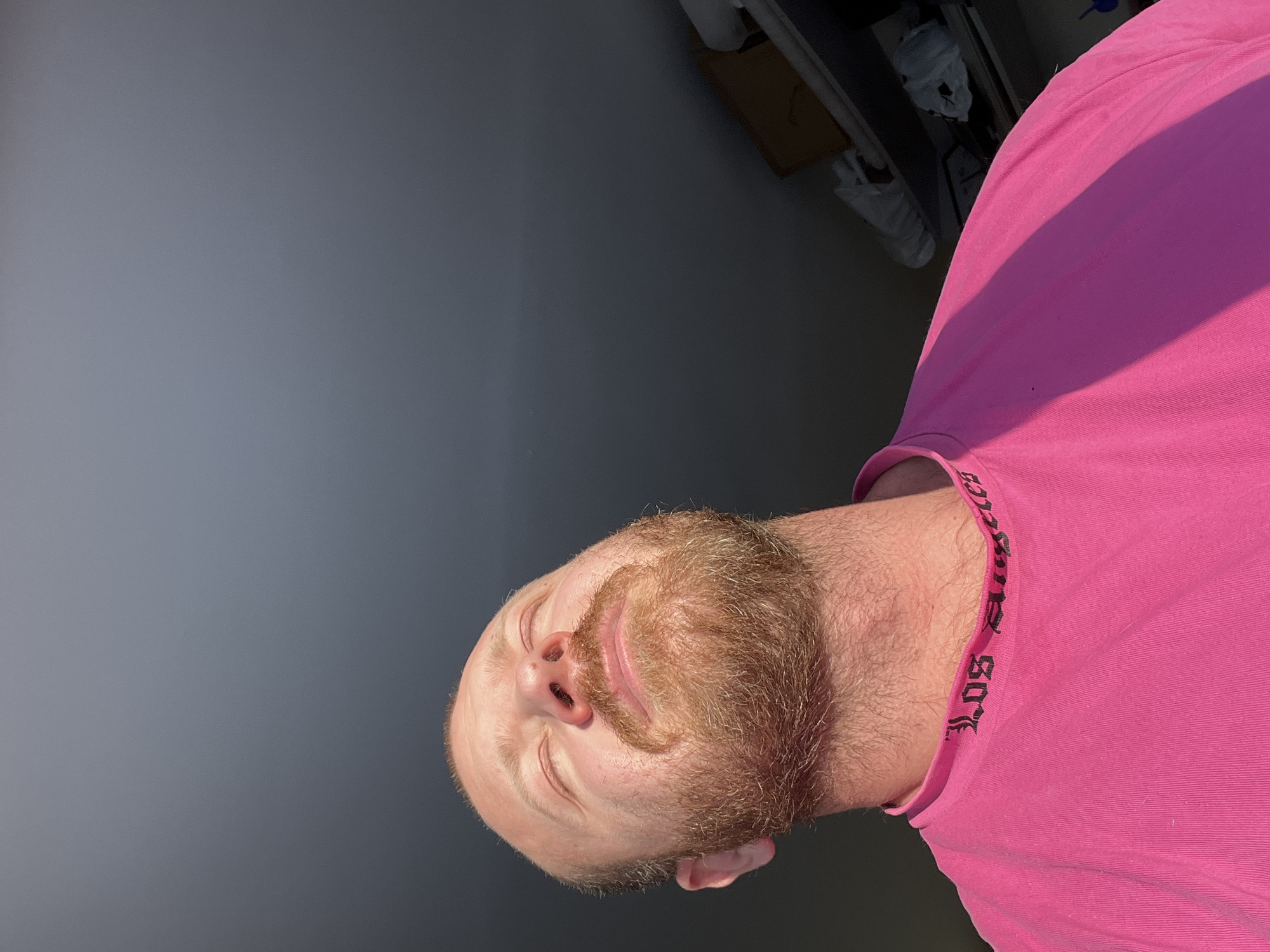 An image taken in the city of Noviy, Russia, depicts a male subject from a low-angle perspective, illuminated by strong natural light from the left. The subject is a fair-skinned male with a balding scalp and a reddish-blonde full beard and mustache. He is looking upwards with his eyes closed, head slightly tilted back. He is wearing a bright pink t-shirt; black text, partially inverted, visible on the collar reads "LOS ANGELES". The left side of his face, neck, and upper torso are brightly lit, while the right side is in shadow. The background consists of a dark grey wall or ceiling. In the upper right corner, an indistinct shelf unit is visible in shadow, holding a brown cardboard box on an upper shelf and several white, possibly plastic-covered, objects on a lower shelf.