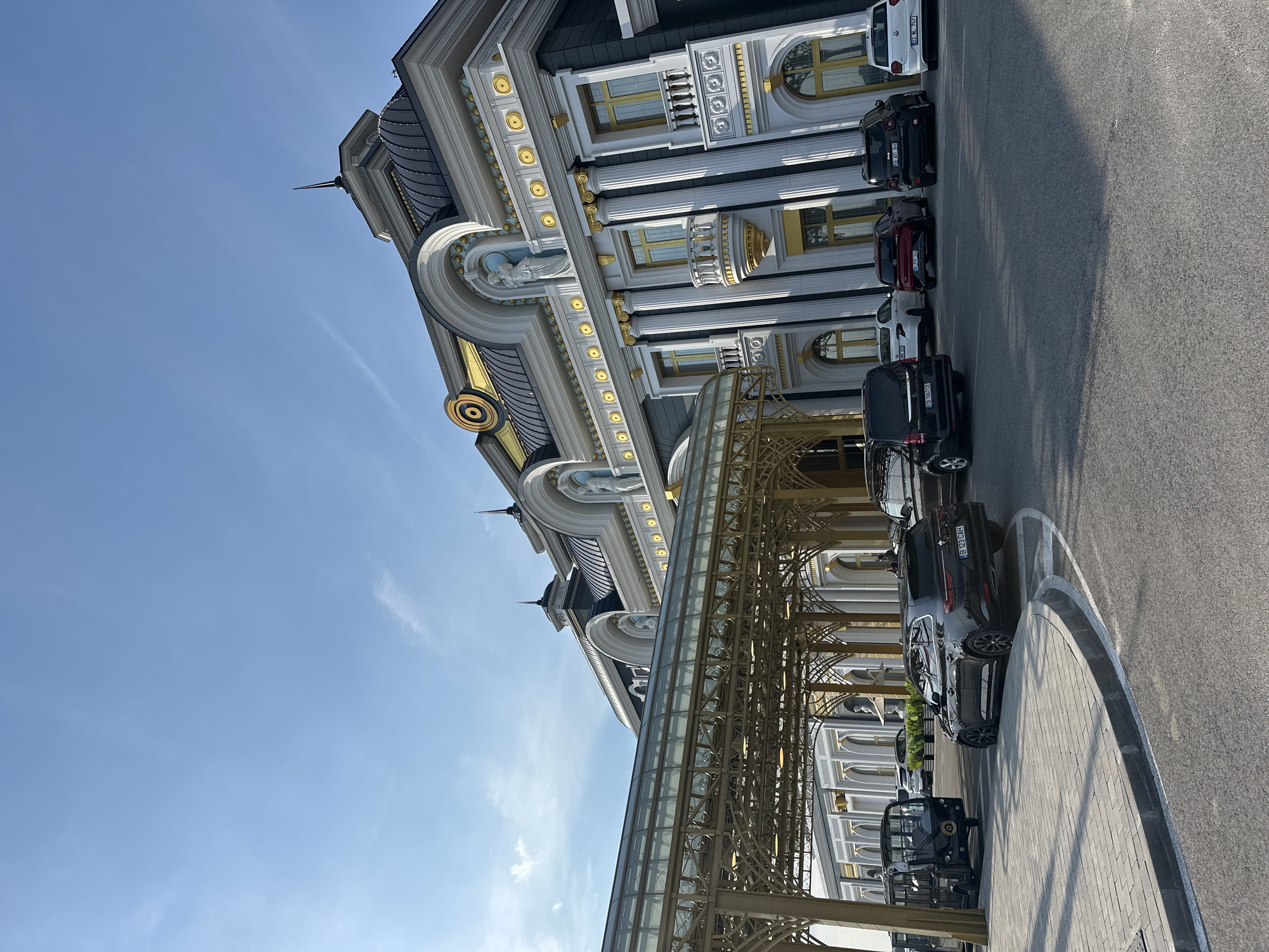 The image displays the exterior of a large, ornate building in Kapitan Andreevo, Bulgaria, under a clear blue sky with scattered cirrus clouds. The building's architecture is characterized by classical and Baroque revival elements, featuring a predominantly white and dark grey facade with extensive gold detailing on cornices, window frames, balustrades, and column capitals. The roofline includes multiple dark-tiled domes and pointed finials. Sculptural figures are visible within arched recesses on the upper levels. A prominent, gold-colored metal and glass canopy extends over a paved driveway, serving as a portico.
Several vehicles are present on the asphalt driveway and parking areas. A black Renault Talisman Grandtour station wagon, bearing Romanian license plate B 123 CAH, is positioned on the driveway, turning. A small, open-sided black utility vehicle is parked near the base of the canopy. Underneath the canopy, a black Mercedes-Benz Vito van with Bulgarian license plate CB 1067 CE is parked. Further along the building's facade, other parked vehicles include a white sedan, a red hatchback, a dark Jeep Renegade with Bulgarian license plate CM 1912 MX, and a white Volkswagen Polo hatchback with license plate 22 ML 747. The ground consists of asphalt for vehicular traffic and lighter grey paver stones for pedestrian pathways and curbs. The scene is illuminated by direct sunlight, casting distinct shadows. Vague human figures are discernible in the distance near the building's entrance.