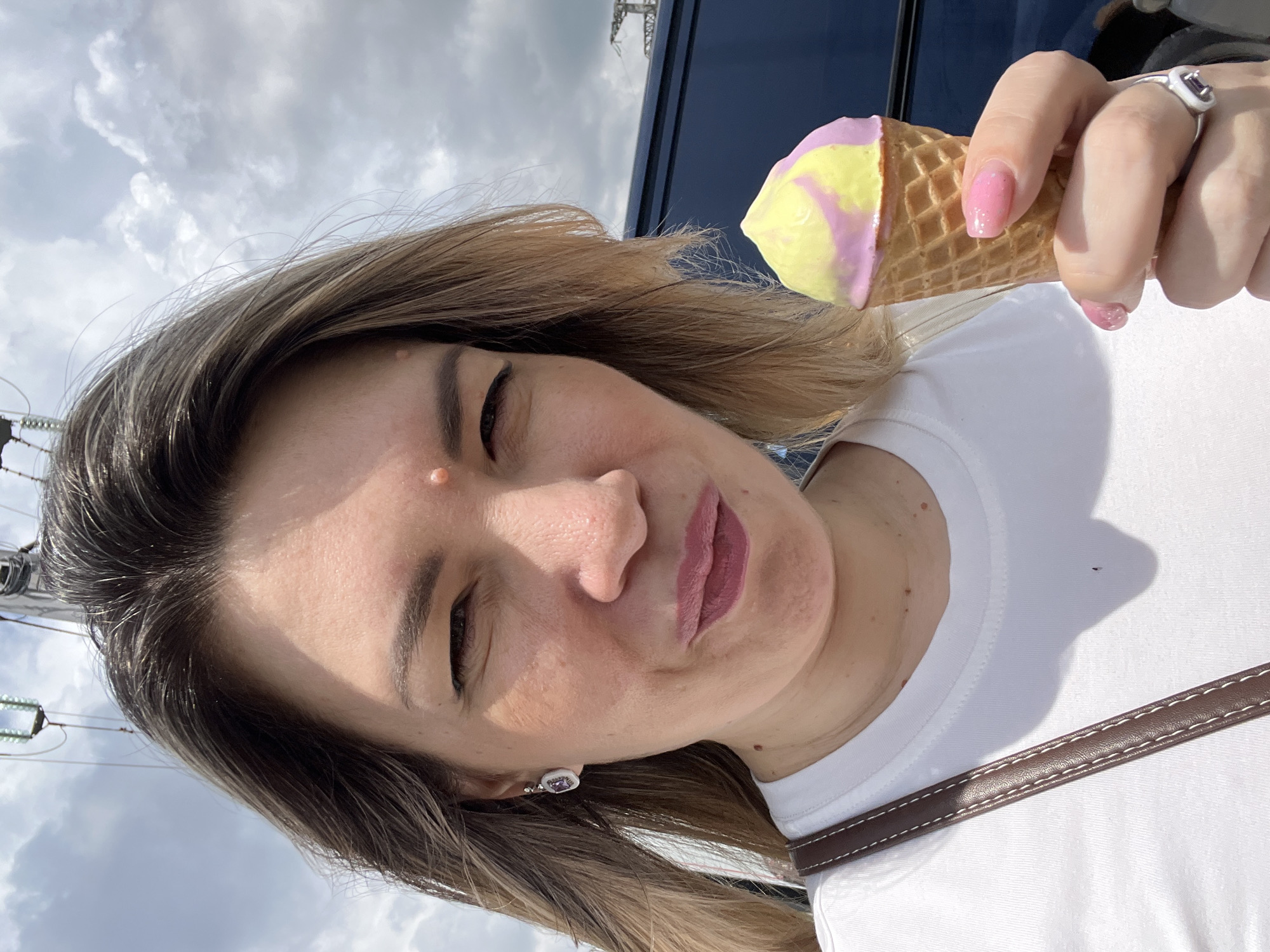 A close-up, selfie-style photograph captures a Caucasian woman with blonde hair and fair skin holding a pink and yellow swirl ice cream cone in her right hand. She is wearing a white t-shirt and a brown strap from a purse. Her expression is one of playful squinting. In the background, the sky is cloudy, and overhead wires are visible, suggesting an outdoor urban setting possibly in Sochi, Russia, given the presence of similar infrastructure near coastal areas. The overall lighting indicates it is daytime.