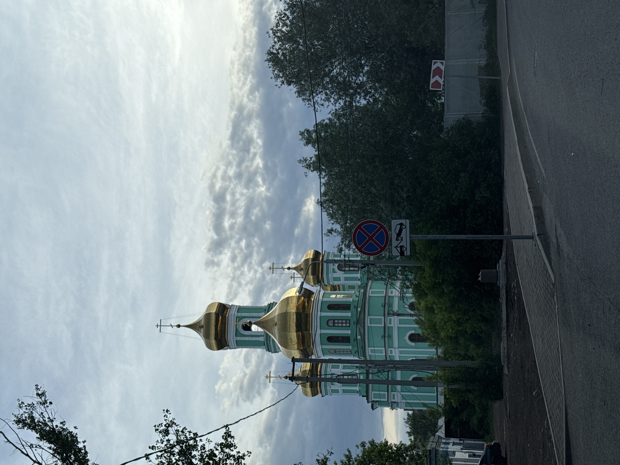 The image shows the Holy Trinity Stefan Monastery in Perm, Russia, from a street-level perspective. The monastery is a green and white building with several golden domes, partially obscured by trees. A "no parking" sign is visible in the foreground, along with a sign indicating a sharp turn. The sky is cloudy, and the road is on the right side of the frame. A power line runs across the bottom of the image.