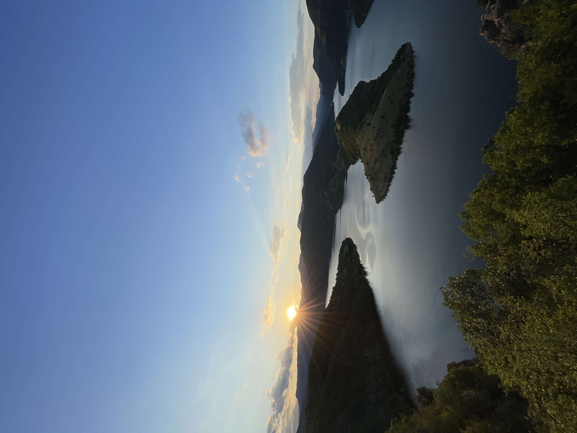 This is an aerial shot of a wide, serpentine river with dark, still water, seen at sunset. Rolling, tree-covered hills surround the river, with a few small, grassy islands jutting out of the water. The sky above is a clear, light blue, with a few wispy clouds tinged pink and orange by the setting sun. The sun itself is low on the horizon, peeking out from behind a cluster of hills and casting long rays of light across the water and sky. The foreground is dominated by the dark green foliage of trees, suggesting the photo was taken from a high vantage point overlooking the landscape. This scene captures the natural beauty of the area surrounding Borovitsa, Bulgaria, specifically the Tsankov Kamak dam reservoir.