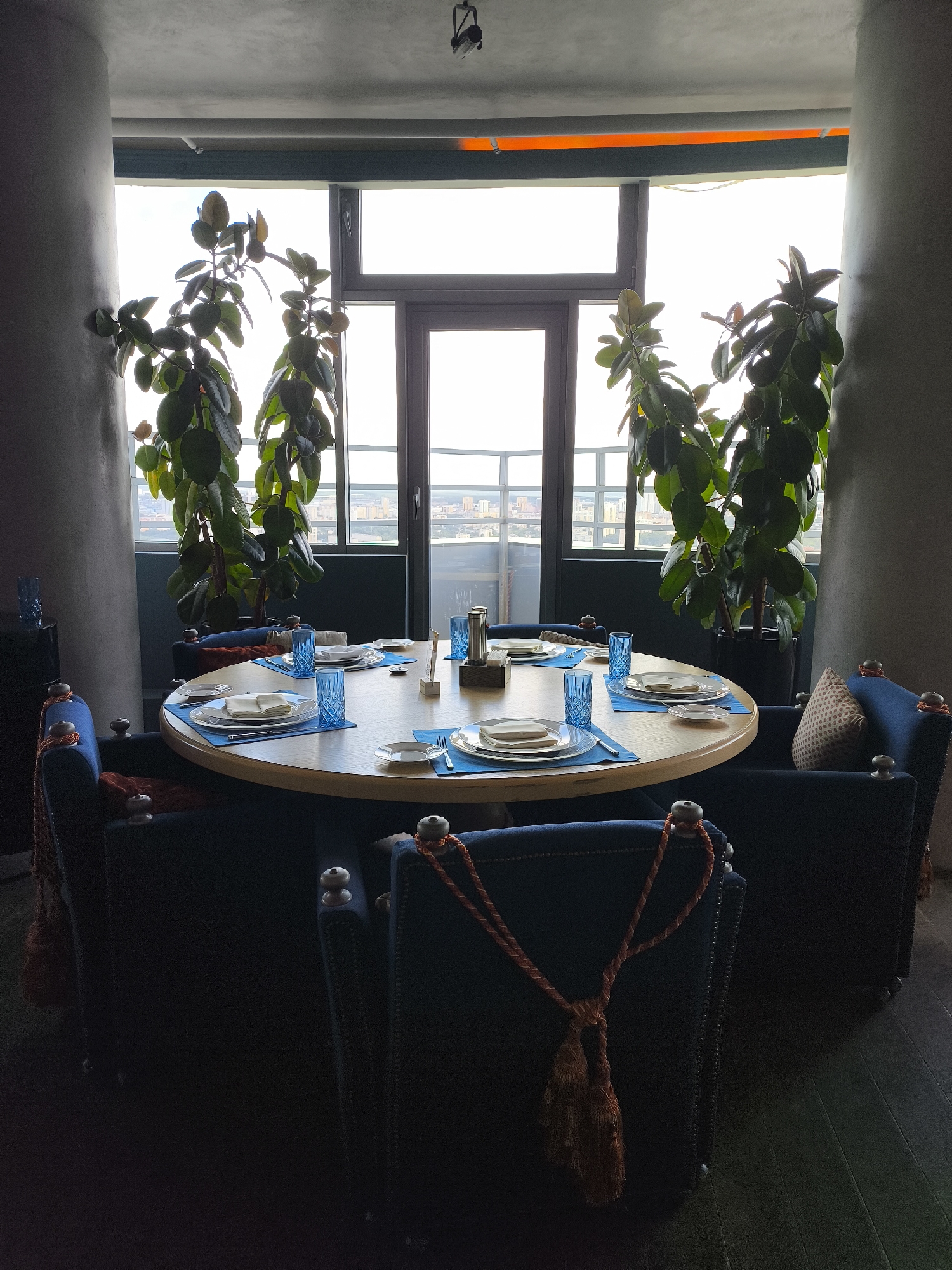 The image depicts a circular dining table set for multiple diners in a dimly lit restaurant. The table is round and made of light wood, with place settings that include white plates, silverware, and blue glassware. Blue placemats and a central wooden object with a metal stand are also visible. The chairs surrounding the table are upholstered in dark blue fabric and feature decorative orange tassels.
Behind the table, a large window offers a view of a city skyline, suggesting an urban environment. Two tall potted plants with broad, green leaves flank the window, adding a touch of nature to the interior. The overall ambiance is sophisticated and slightly moody, with the light filtering through the window and casting shadows within the room. The presence of the table set for dining indicates a restaurant setting, and the view of the skyline suggests it is located in a city. The specific city of Yekaterinburg, Russia, is not explicitly identifiable from the visual cues alone; however, if this were a known location, the description would be the same, focusing on the visual elements present.