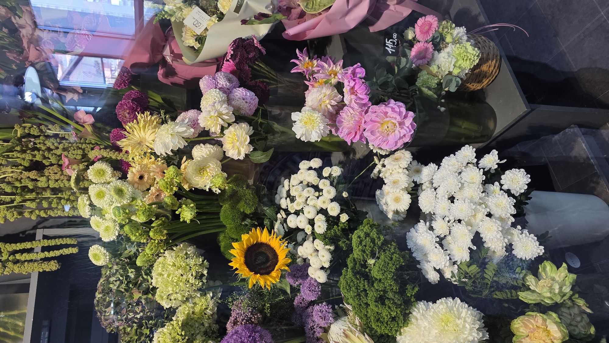 A close-up, eye-level, outdoor shot displays a variety of fresh flowers arranged in what appears to be a floral shop or market stall. The dominant colors are white, pink, yellow, and green, with varying textures and bloom shapes. In the center, a bright yellow sunflower with a dark brown center stands out against a backdrop of white chrysanthemums and other light-colored flowers. To the left, a large, textured green bloom is visible, along with lighter green hydrangeas. Pinks and purples are present in smaller quantities, seen in dahlias and other filler flowers. The flowers are arranged in vases, baskets, and directly on display. Reflections from a window are visible in the upper right portion of the image, showing a hint of buildings and sky, suggesting an urban setting. A price tag reading "115,00" is visible on the lower right. While the image is focused on the floral display, the reflections suggest the location is Brest, Belarus, possibly a shop on a street with reflections of buildings typical of the city. No people are visible in the frame, and no actions or interactions are depicted.