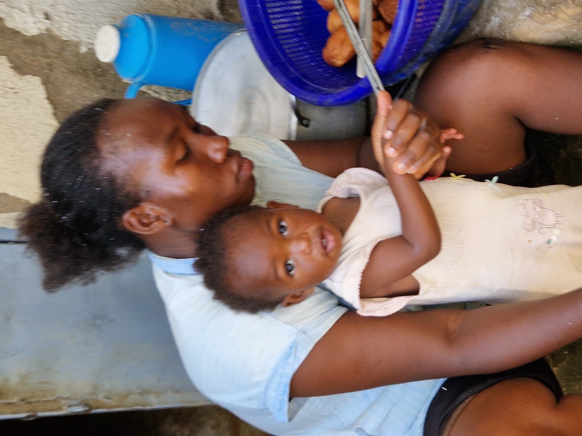 In a dimly lit outdoor setting, possibly in the city of Jos, Nigeria, a dark-skinned woman with her hair pulled up in a bun is seated, holding a baby. The woman is wearing a light blue short-sleeved shirt and black shorts. The baby, also dark-skinned, is wearing a light-colored, possibly tan, onesie with some embroidered details and is facing forward. The woman's arms are around the baby, and her head is slightly bowed, looking down.
To the right of the woman and baby, a bright blue plastic basket is filled with what appear to be fried dough balls. A pair of metal tongs is resting on the rim of the basket, with one end submerged in the fried food. Behind the basket, a large, light-colored pot with a handle is visible. Further in the background, a bright blue plastic container with a white lid, resembling a thermos or water jug, stands upright against a rough, textured wall. The ground beneath them appears to be made of rough, textured concrete or stone. The overall lighting suggests it is daytime, but the area might be shaded or covered.