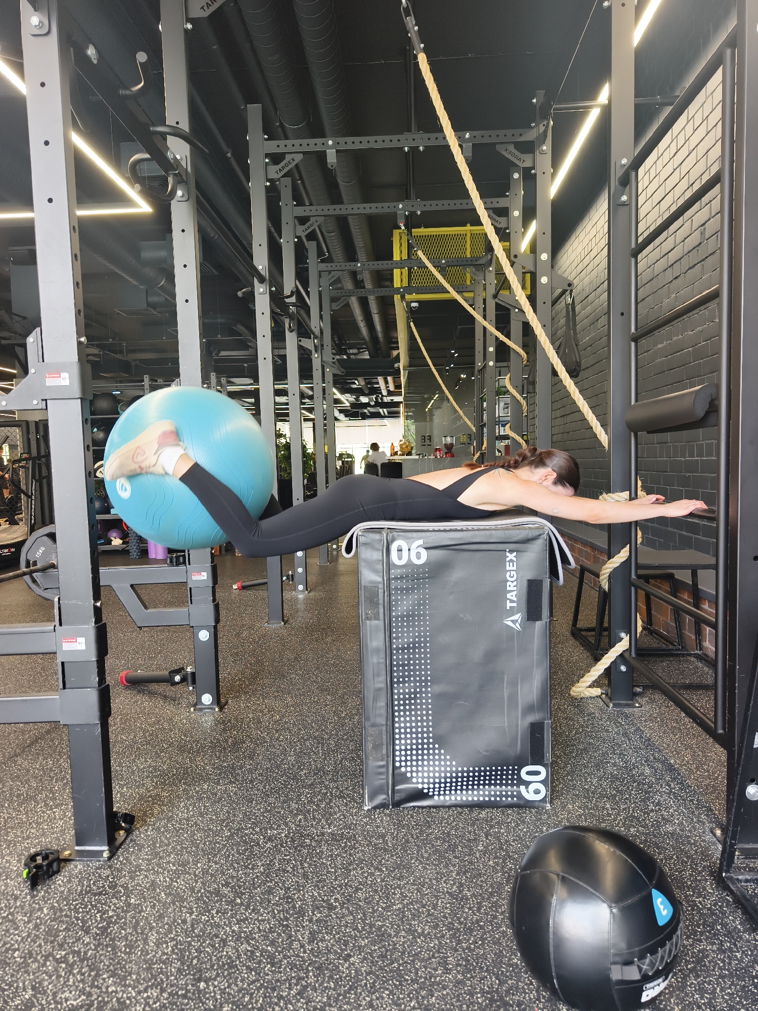 The image displays a female athlete engaged in a fitness exercise within a modern gym setting, possibly located in Tyumen, Russia. The woman, dressed in a black athletic bodysuit and leggings with her dark hair pulled back, is positioned prone across a large black plyometric box. Her legs are elevated, firmly holding a large blue stability ball between her calves and feet. Her upper body extends horizontally forward, with her arms stretched out in front of her. Her face is not visible, but her pose indicates focused effort.
The gym environment features a robust black metal training rig with various bars and hooks, suggesting a functional training or CrossFit facility. Several thick beige ropes hang from the ceiling. A black painted brick wall is visible on the right side of the frame. The floor is covered with speckled black rubberized gym matting. The ceiling reveals exposed ventilation pipes and bright linear strip lighting, contributing to an industrial aesthetic.
Visible textual details include "TARGETX" printed on the side of the black plyometric box and also on parts of the gym rigging, indicating the brand of the equipment. The box additionally shows the numbers "06" and "60," with "60" likely referring to its height in centimeters. A black medicine ball with a blue logo rests on the floor. Part of a weight plate is visible, marked "15KG." The bright lighting suggests it is daytime.