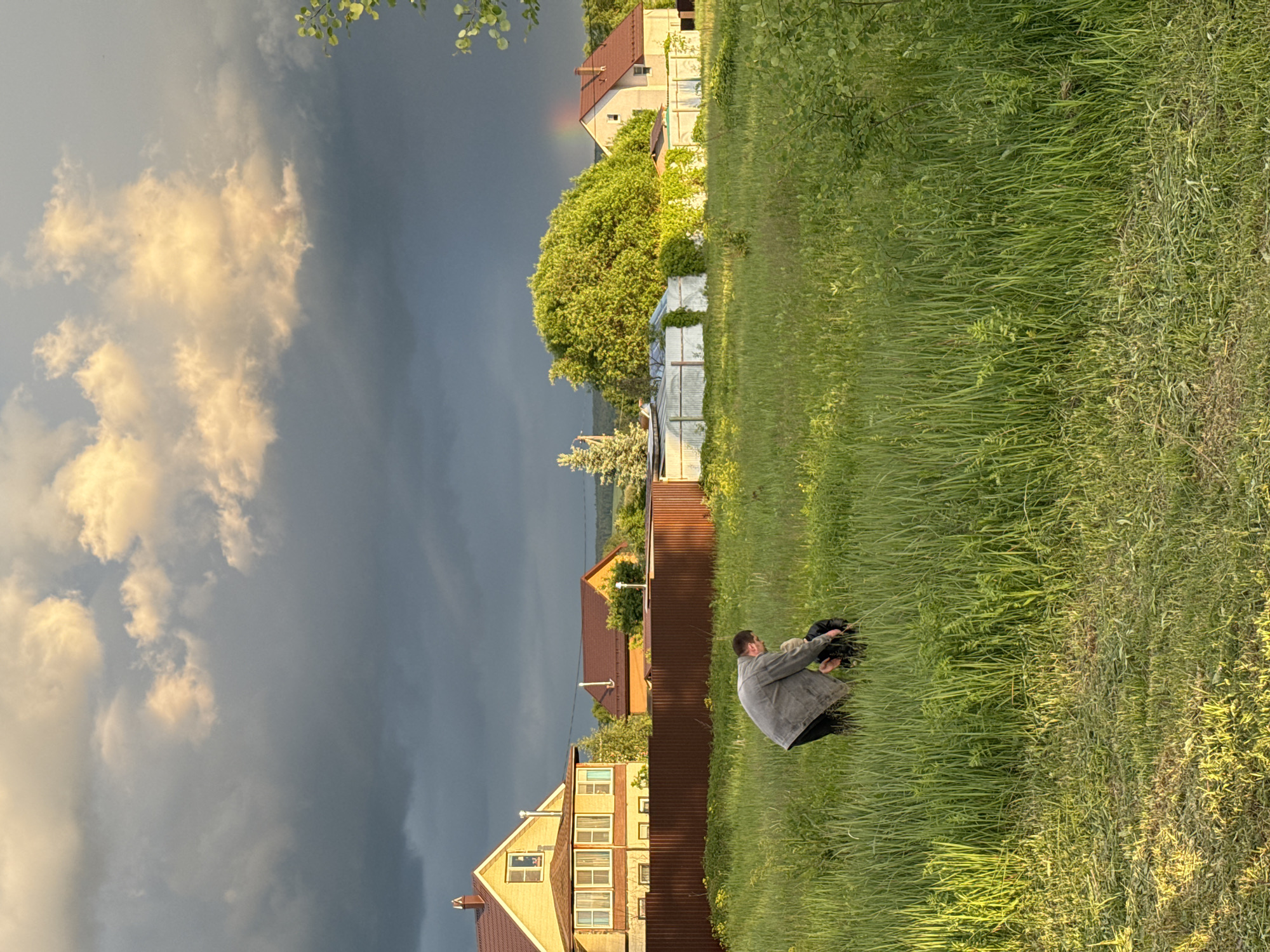 A medium, landscape-oriented shot shows a man kneeling in a grassy field in Vysokinichi, Russia. Storm clouds fill the left sky, while residential houses line the horizon.
The man, dressed in a grey jacket and black pants, is kneeling in the middle-right of the frame. He holds something dark in his hands.
Behind him, a brown fence separates the field from a row of houses. The houses are varied in color and style, with one being yellow and another having a red roof.
The field is covered in tall green grass, with patches of lighter and darker shades. The sky is filled with dramatic storm clouds on the left, contrasting with the sunlight on the right. Some trees can be seen in the background, along with a few other buildings.