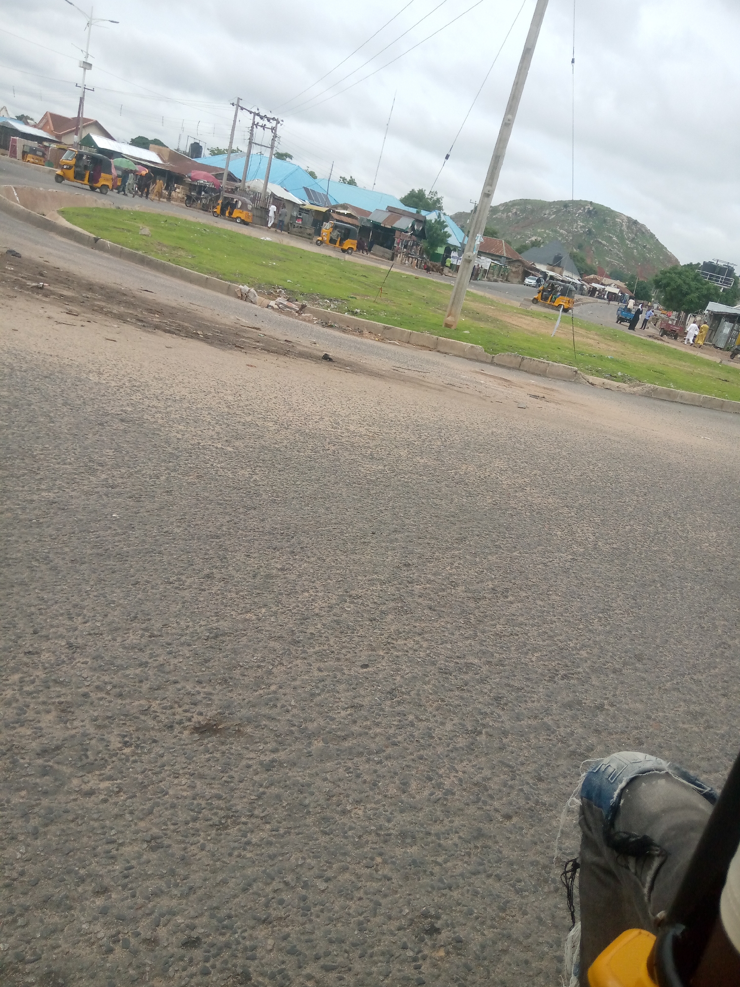 The image depicts a street scene in Jalingo, Nigeria, with a focus on a busy intersection. In the foreground, a portion of a vehicle's seat and a person's leg wearing torn jeans are visible, suggesting the photo was taken from inside a vehicle. The road surface is asphalt with visible gravel. Across the street, a market area is set up with various stalls and shops. Several "Keke Napep" (three-wheeled motorized rickshaws) are parked and in motion, indicative of local transportation. People are seen walking, some carrying umbrellas, and interacting in the market. The background features a prominent rocky hill, a characteristic geographical feature of Jalingo. Electrical poles and wires are present, common in urban environments. The sky is overcast, suggesting a cloudy day. The overall scene portrays a lively commercial area with typical elements of Nigerian street life.