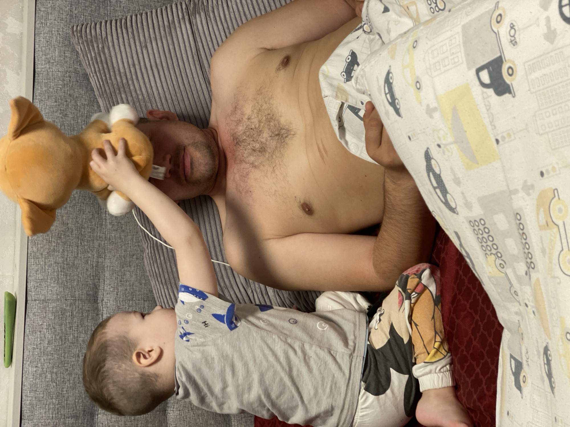 This indoor photograph, taken in Sochi, Russia, depicts an adult man and a young child on a bed. The man is lying shirtless on his back on a grey striped sheet, with his eyes closed, suggesting he is resting or asleep. A white medical tube is visible near his right nostril and extends towards his mouth, indicating a potential medical need or recovery.
To the man's left, a young child is positioned on the bed, wearing a grey t-shirt with blue patterns and white pajama bottoms with Mickey Mouse designs. The child is holding an orange plush dog toy and reaches towards the man's face with it, seemingly engaged in play. The child's t-shirt has the word "HI" printed on it.
Part of the bed is covered by a light-colored blanket featuring a pattern of cars and buildings. The word "STOP" is visible on one of the car designs, and multiple "000" markings are present. This blanket partially covers the man's right arm and lower body. A dark red patterned sheet is also visible beneath the car-patterned blanket. The scene captures a quiet, intimate moment between the two individuals.