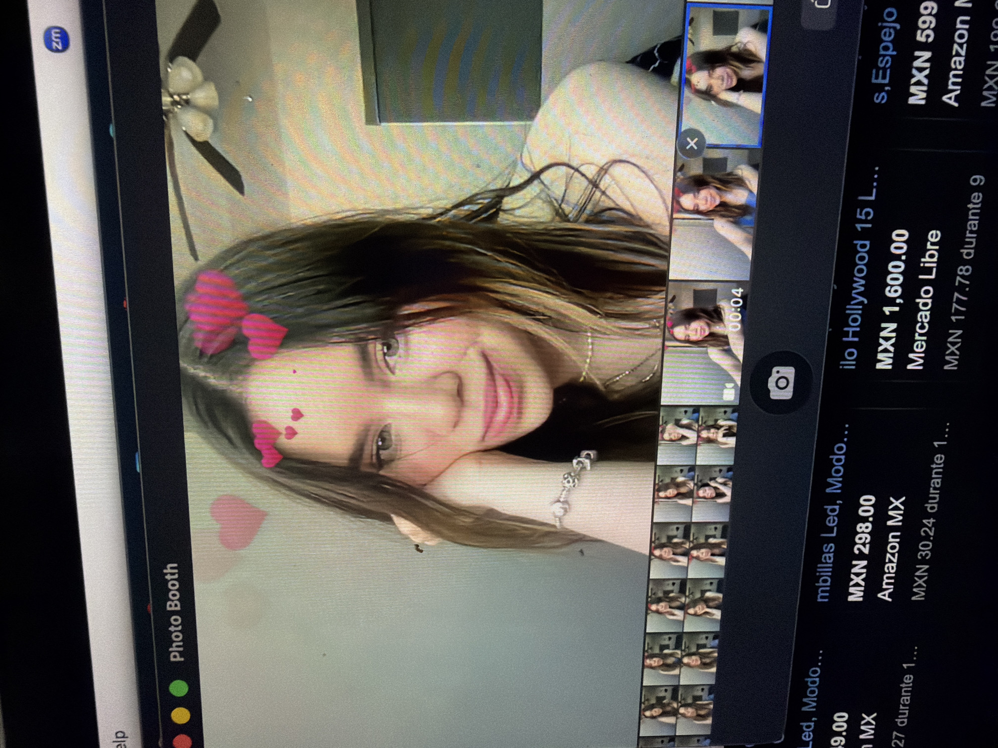 The image displays a photograph of a computer screen, presumably located in Tuxtepec, Mexico. The screen shows two distinct applications: "Photo Booth" in the upper portion and a web browser displaying e-commerce listings in the lower portion.
Within the "Photo Booth" application, a young female subject is visible, smiling subtly and resting her left hand against her cheek. She has long brown hair with lighter streaks and wears a delicate necklace and a silver-colored charm bracelet on her left wrist. Digital pink heart overlays appear around her head, indicating the use of a filter. The background behind the subject is a light-colored wall with a dark-bladed ceiling fan and a light fixture visible in the upper right. Below the main Photo Booth display, a grid of smaller thumbnail images shows the same female subject in various poses, some with additional digital accessories like red horn-like headbands. A camera icon is present in this section.
Below the Photo Booth interface, a partial view of a web browser reveals product listings from Mexican e-commerce platforms. Visible text includes "mbillas Led, Modo...", "MXN 298.00" from "Amazon MX", and "ilo Hollywood 15 L..." priced at "MXN 1,600.00" from "Mercado Libre," indicating transactions in Mexican Pesos (MXN). The overall scene depicts a user engaged in both casual webcam photography and online shopping on a personal computer within an indoor environment.