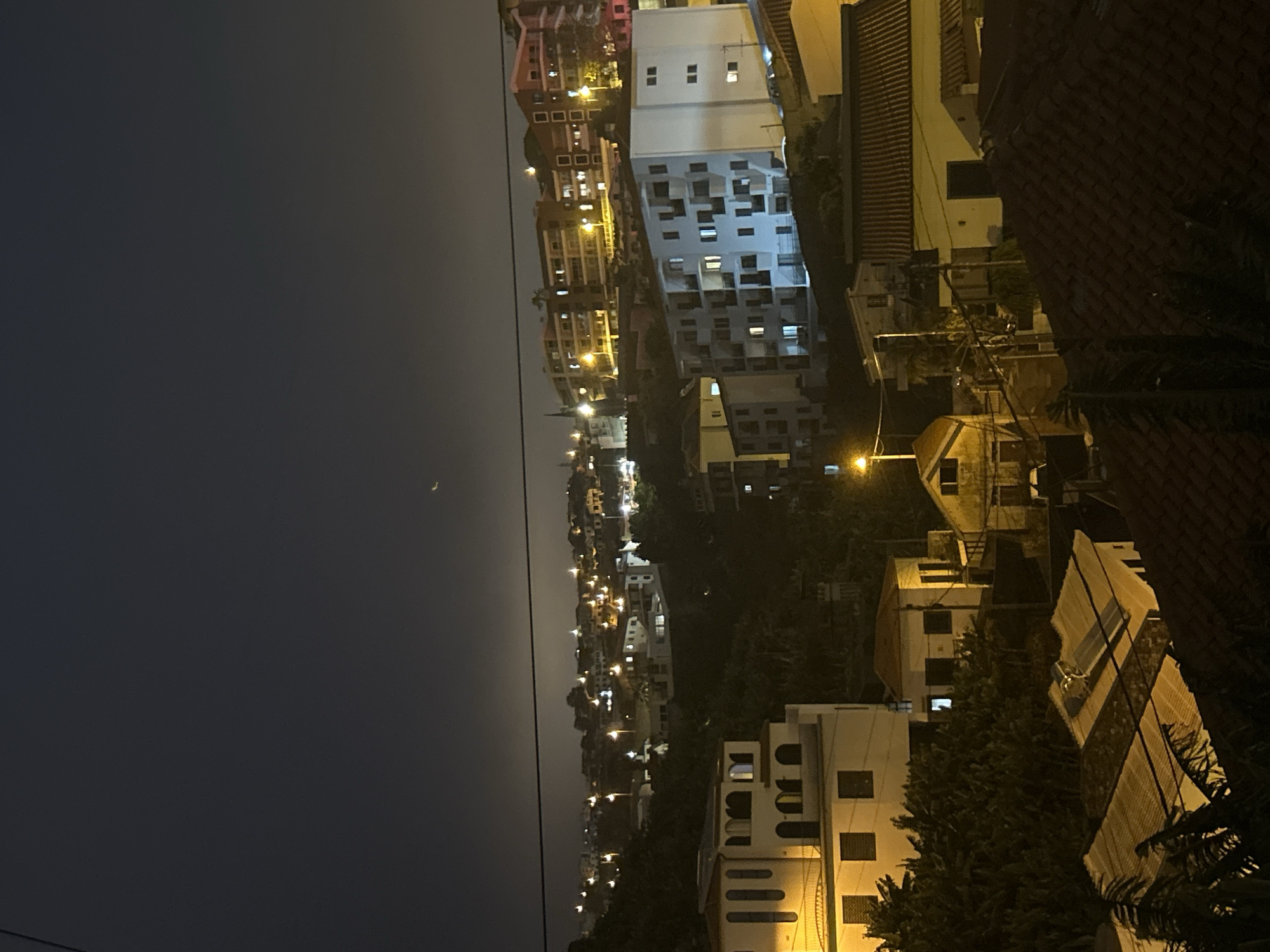 The image presents a night view of Câmara de Lobos, Portugal. The sky, largely dark, reveals a crescent moon. Buildings, predominantly white and beige with lit windows, are seen down the slope. Vegetation and a utility pole are visible.