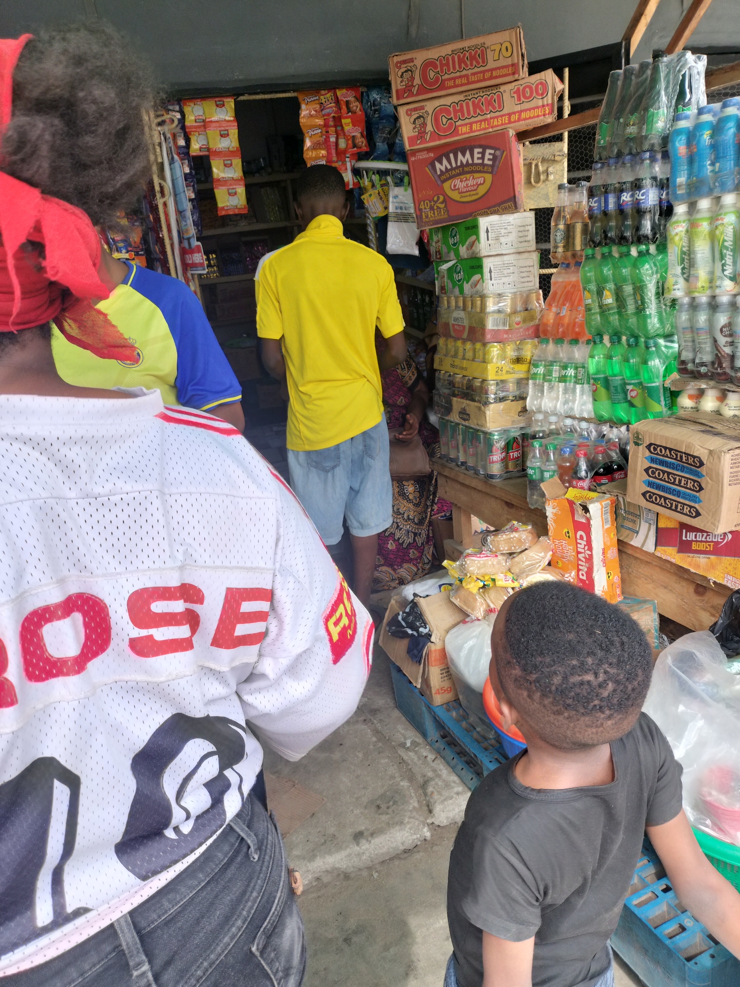 A daytime shot shows the interior of a small store in Alaiyabiagba, Nigeria. Several people are inside, including a child in a black shirt in the foreground, and others looking at merchandise in the back. The shelves are stocked with a variety of items, including instant noodles (Chikki 70, Chikki 100, Mimee), bottled drinks, canned goods, and snacks. A white sports jersey with "OSE" in red block letters is visible in the lower left.