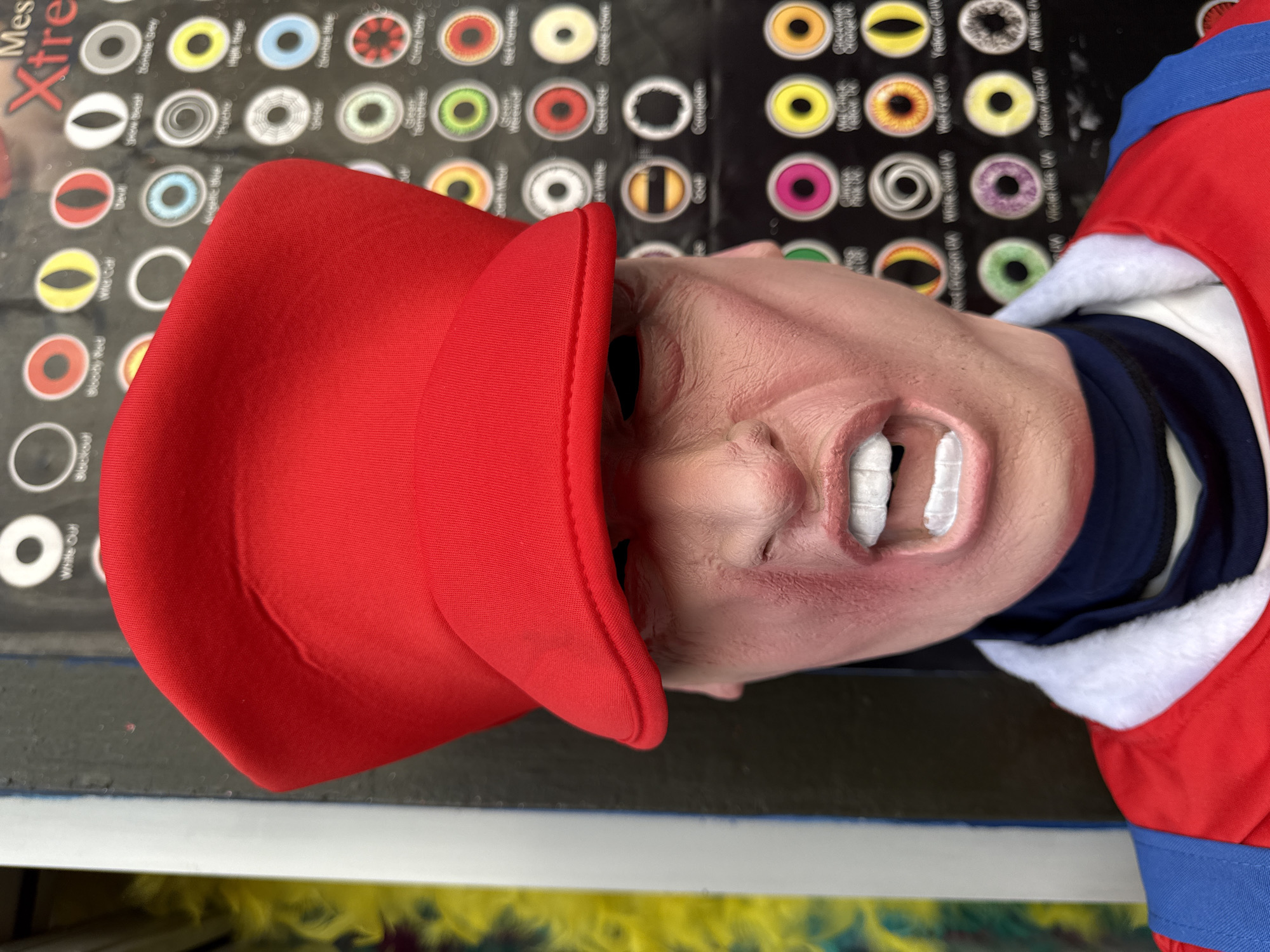 A close-up shot features a rubber mask of a person wearing a red cap and a red and white garment. The mask's mouth is open, revealing prominent white teeth. The face is light-skinned with wrinkles and fine details. The mask appears to be placed against a backdrop with a poster displaying various eye designs, labeled with names such as "Devil," "Wild Cat," "Zombie Blue," and "Creepy Baby." The text "Mes Xtre" is visible on the left side of the poster. The scene is set on a surface, with a blurred lower section suggesting it's on a shelf or counter. The absence of street view imagery prohibits determining if the photograph was taken in London, United Kingdom.
