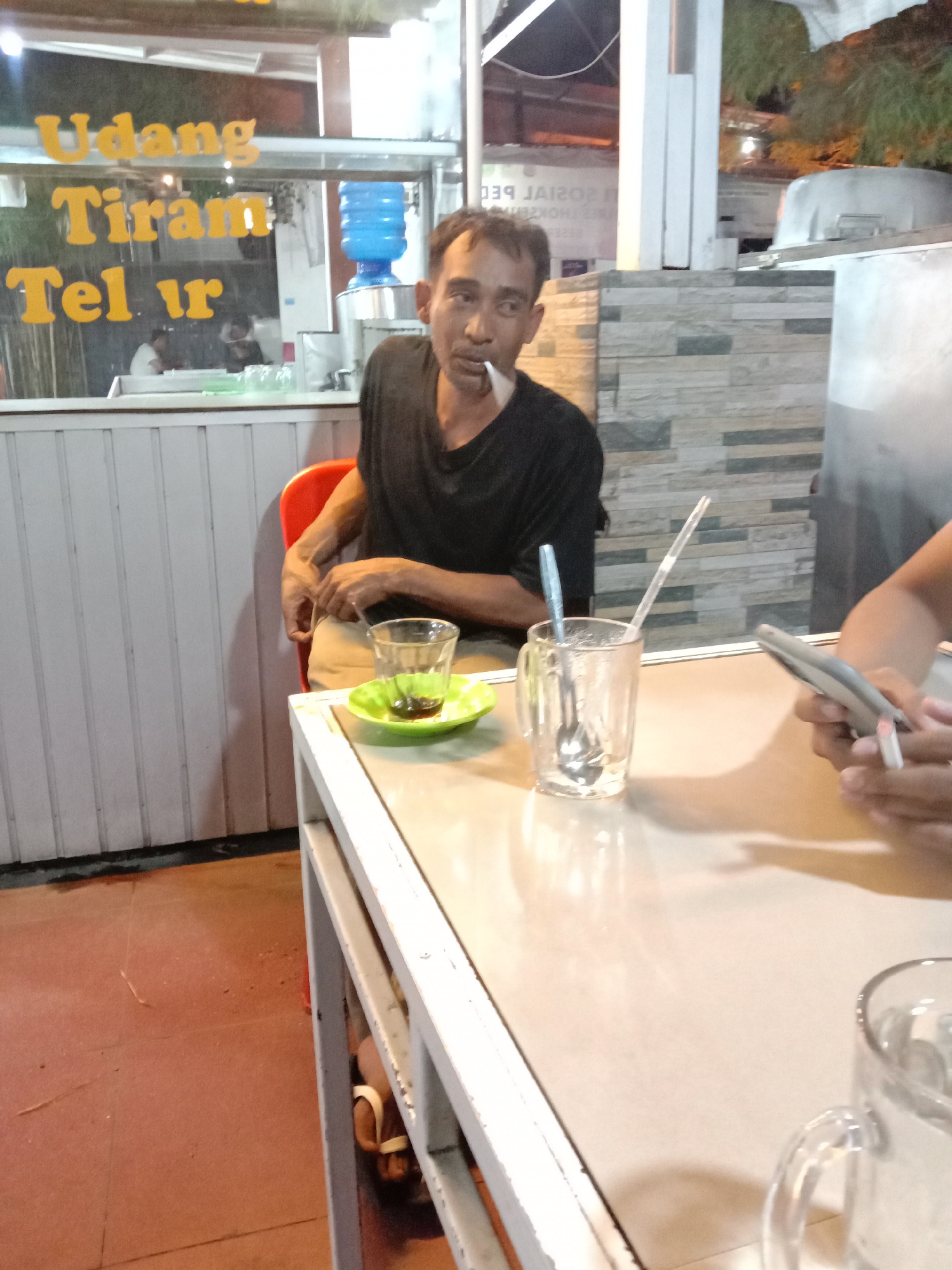 The image depicts an indoor scene at night, likely a restaurant, in Lhokseumawe, Indonesia. In the foreground, a white metal table is visible, reflecting the ambient light. On the table, there are several objects: a glass mug with a spoon and straw, a small glass with a green saucer containing a dark liquid, and a hand holding a mobile phone. In the mid-ground, a man is seated on a red chair. He is wearing a black t-shirt and appears to be smoking a cigarette. Behind him, a water dispenser and a tiled wall are visible. Above the man, text in yellow font on a glass window reads "Udang Tiram Telur". The overall lighting suggests an artificial, evening setting.