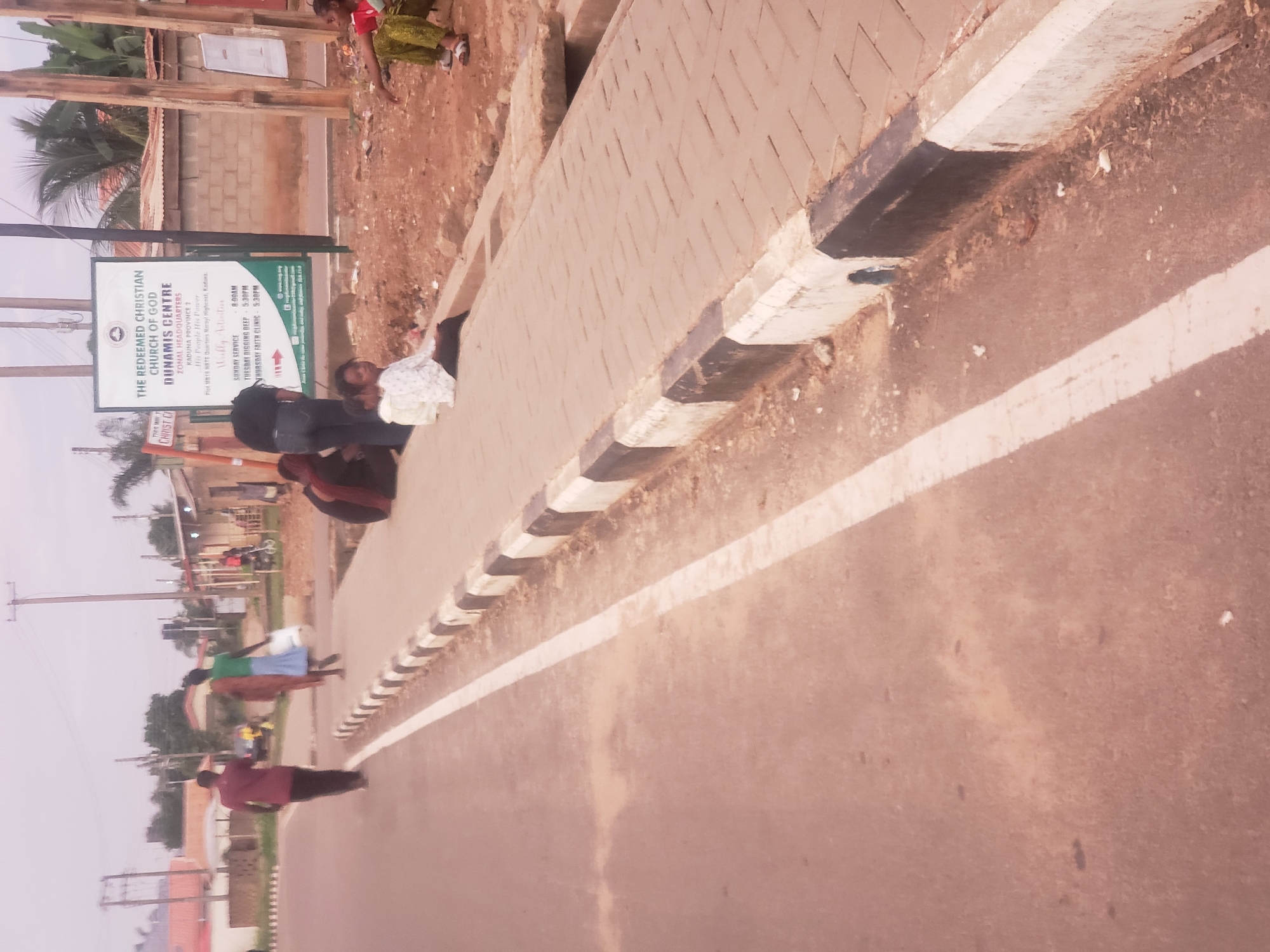 The image depicts a street scene in Narayi, Nigeria. In the foreground, a paved road with a white lane marker runs parallel to a sidewalk constructed of interlocking concrete pavers. The sidewalk is bordered by a curb painted with alternating black and white bands.
Several individuals are present. On the sidewalk, a person in a white and black patterned shirt and dark pants is seated, facing towards the viewer. To their left, another person, partially obscured, is also seated. Further to the left, two figures are walking away from the camera on the road. One, in a maroon shirt and dark pants, is ahead of the other, who is wearing a green top and a long brown skirt or fabric, carrying a white plastic bag.
In the background, a large white sign with text and a logo is mounted on poles. The sign reads "THE REDEEMED CHRISTIAN CHURCH OF GOD DUNAMIS CENTRE" and lists various "Weekly Activities" and contact information. Buildings with corrugated metal roofs are visible behind the sign, as well as trees and utility poles. A dirt area with some debris is located between the sidewalk and the background structures. The sky is overcast.