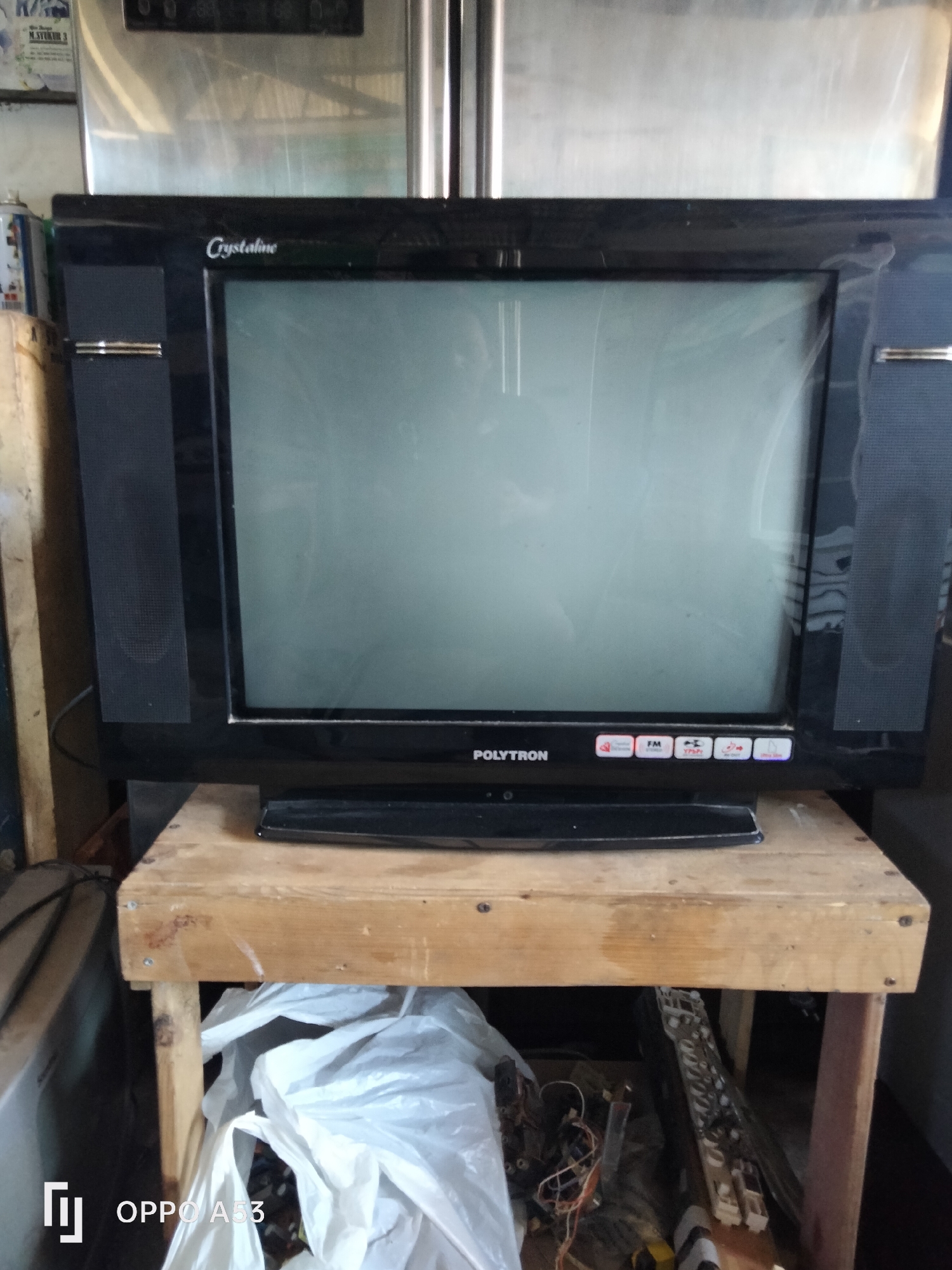 The image shows a vintage television set, identified by the branding "POLYTRON" and "Crystalline" on its frame, resting on a wooden stand. The television is black with visible speakers on either side of the screen, which is currently displaying a light gray image, suggesting it is powered on or in standby mode. Below the screen, there are several small icons and the text "POLYTRON". The wooden stand appears to be a makeshift structure, possibly constructed from reclaimed wood, with visible screws. Underneath the stand, a white plastic bag filled with various items, including electrical wires and components, is visible. The background is somewhat obscured but suggests an indoor environment with what appears to be a large appliance, possibly a refrigerator, behind the television. The image also contains a watermark in the bottom left corner that reads "OPPO A53". The overall scene depicts a cluttered setting, typical of a storage area or a less organized living space. Without additional context or clues, pinpointing the exact location in Denpasar, Indonesia is not possible from the visual information alone.