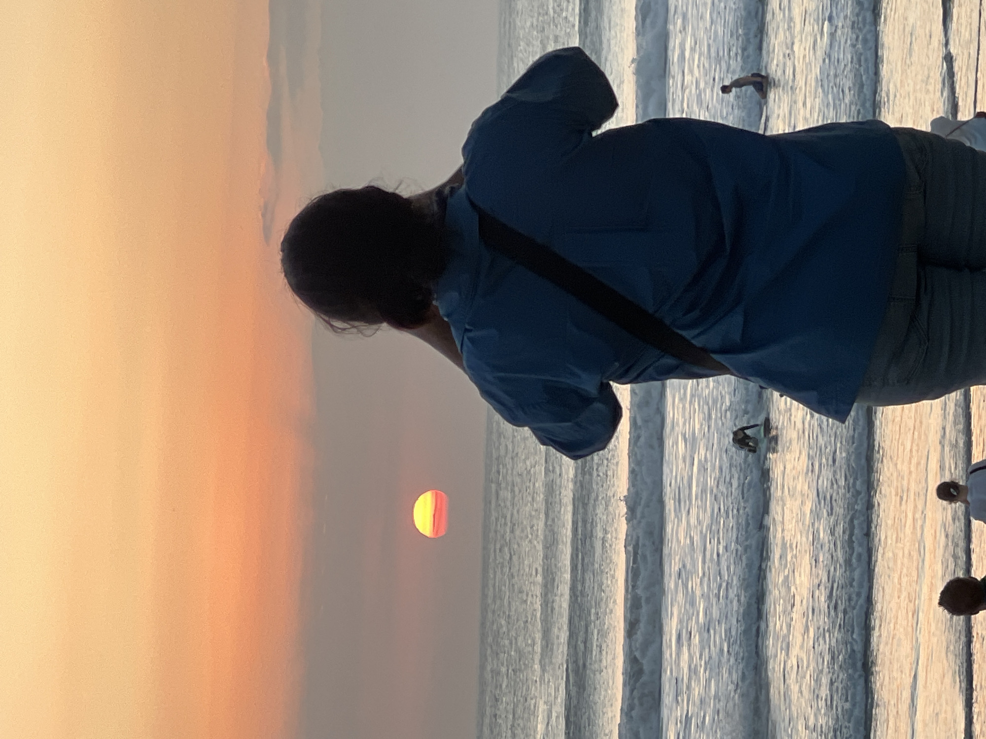 The image captures a sunset at Kuta Beach, Bali, Indonesia. The sun, a vibrant orange and yellow orb, is low on the horizon, casting a warm glow across the sky and reflecting on the gentle waves. The ocean is a mix of blues and grays, with white foam cresting on incoming waves.
In the foreground, a person wearing a blue shirt and gray pants stands with their back to the camera, holding a phone up to take a picture of the sunset. A dark strap from a bag is visible across their shoulder.
Further out in the water, several surfers are visible. One surfer is kneeling on their board, while another is further out, partially submerged in the water. Two other individuals are visible in the background, standing on the shore. The overall atmosphere is serene and peaceful, with the natural beauty of the sunset dominating the scene.