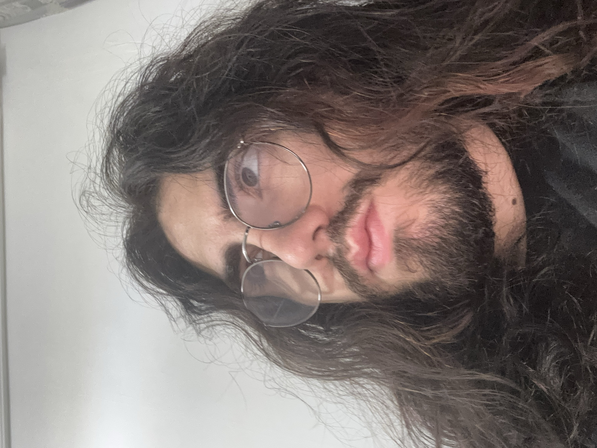 The image is a close-up, low-angle shot of a person with long, dark, wavy hair and a beard. They are wearing round, wire-rimmed glasses and looking upwards and to the left. The person's skin is light-toned. The background is a plain, light-colored wall, possibly a ceiling or a bright wall. There are no identifiable landmarks or features to suggest a specific location within a city. The lighting appears to be artificial and diffuse.