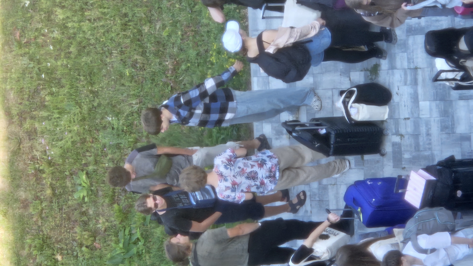 In an overhead shot, a group of young people is gathered on a paved area with grassy terrain behind them, likely at the bus station in Prešov, Slovakia. The individuals appear to be preparing for or concluding travel, as evidenced by the luggage scattered around them.
Several people are standing and conversing. One young man, wearing sunglasses and a black t-shirt with a graphic, stands to the left. Next to him, a person in a grey t-shirt and a green fanny pack is visible. To the right of them, a person in a blue and black plaid shirt and light blue jeans talks with others. In the foreground, several individuals are more clearly visible. A person in a floral print shirt and khaki pants is standing and looking down. To their left, a person in a grey shirt and black pants holds a white bag. A person in a light pink top and denim shorts, wearing a white cap and a black backpack, stands further right.
The luggage includes a bright blue suitcase, several black rolling suitcases, and various duffel bags and tote bags. One pink box is stacked on top of a black suitcase. The paved ground is made of large, rectangular grey tiles. The background consists of a grassy slope with some scattered small plants and flowers. The lighting suggests it is daytime, possibly late morning or early afternoon.