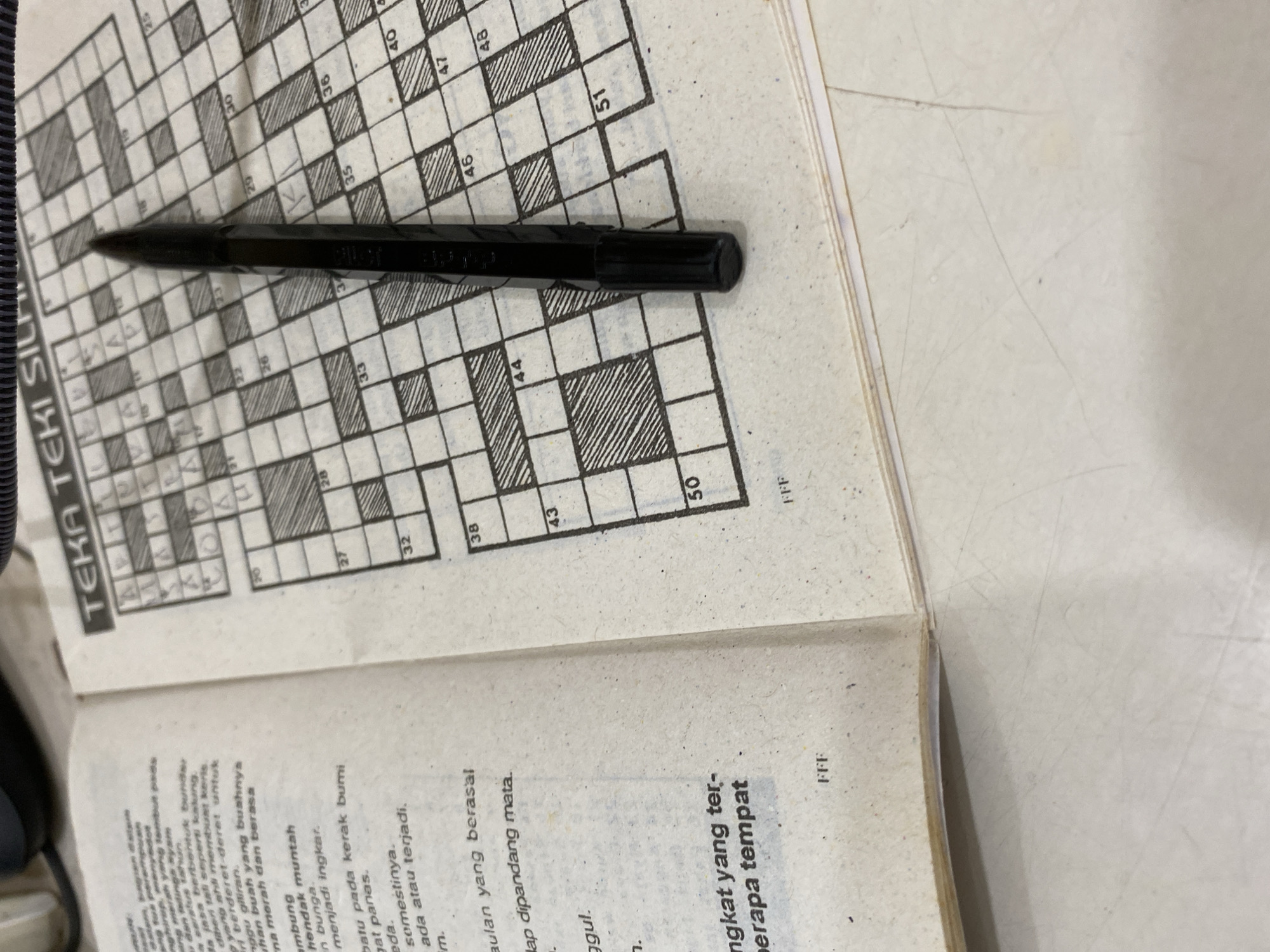 An open book lies on a flat surface, with the right page displaying a crossword puzzle titled "TEKA TEKI SILANG". A black hexagonal pencil with a sharpened tip is positioned diagonally across the crossword grid, its tip pointing towards the top right. The book's left page contains text in Indonesian. The surrounding surface appears to be a light-colored desk or table, with some faint scratches visible. The lighting suggests it is an indoor environment, possibly a desk in a home or office.