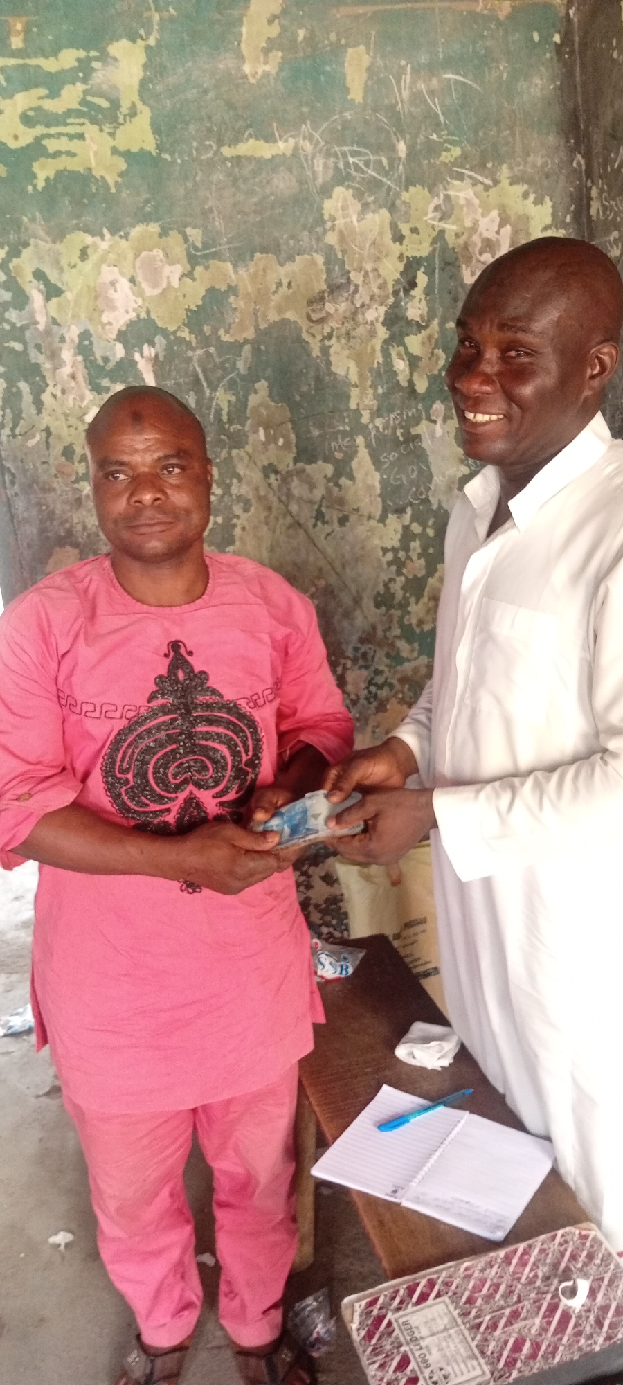 Two men are in an indoor setting. The man on the left wears a pink outfit with a patterned front and is holding money. The man on the right, in a white garment, hands the money to the man on the left. A notebook and pen are on a table to the right, along with a ledger. The wall in the background shows signs of wear and has writing. Location is Unknown, Unknown.