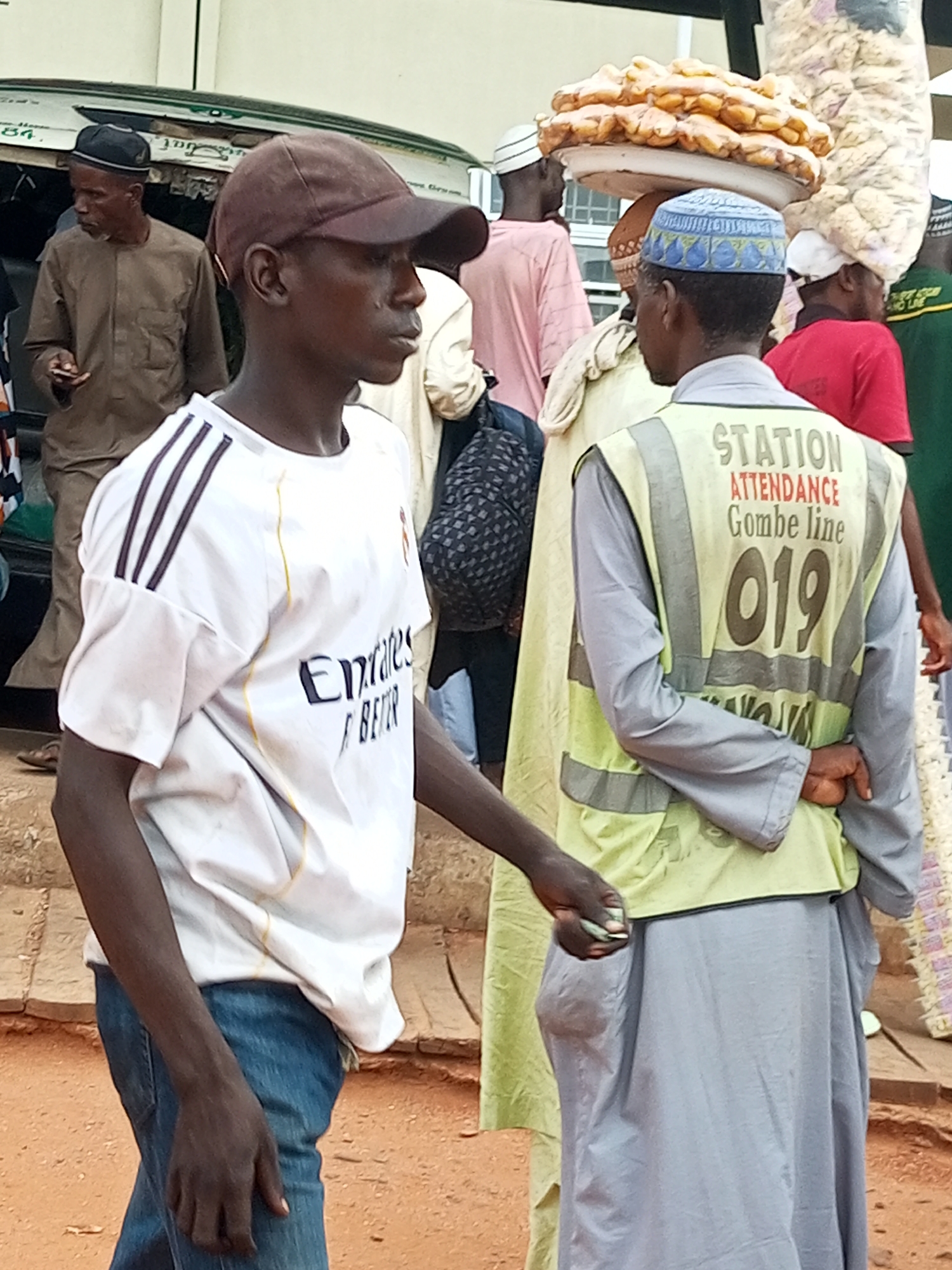 In Gombe, Nigeria, an outdoor scene depicts a group of people, possibly near a transportation hub or market. In the foreground, a young man wearing a brown baseball cap and a white t-shirt with dark stripes on the sleeves and "Emirates" printed on the chest, is partially visible from the side. He is dressed in blue jeans. To his right, another individual wears a light green high-visibility vest over a grey garment, with "STATION ATTENDANCE Gombe line 019" printed on the back of the vest. This person is holding something small in their hand. In the background, several other individuals are present. One man wears a traditional brown robe and a black cap. Another person wears a pink shirt, and a woman is visible wearing a red shirt. A tray of food, possibly bread rolls, is balanced on top of someone's head, and a large bag of what appears to be snacks or goods is also being carried. The ground is covered in reddish-brown dirt and some paving. A vehicle with writing on its side is partially visible on the left.