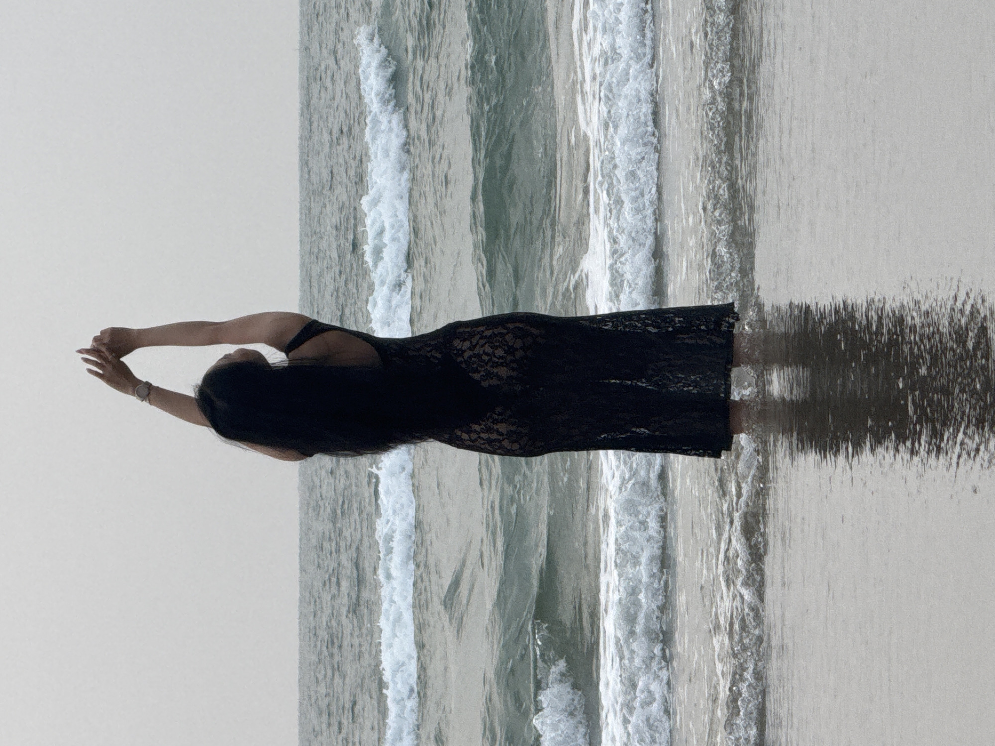 A woman in a black dress stands in the surf on a beach. She is facing away from the camera, with her arms raised above her head. The waves are gentle, and the sky is overcast. The specific location within the city of Đà Nẵng, Vietnam, cannot be determined from the image.