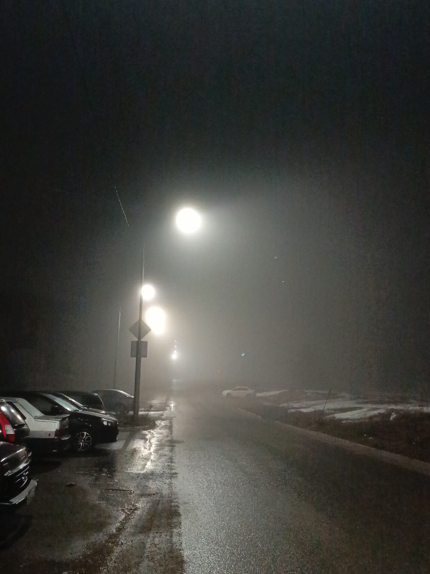 A nighttime scene shows a wet road and sidewalk lined with parked cars in the city of Марковский, Russia. Streetlights cast a hazy glow through dense fog, reducing visibility. A road sign is visible near the parked cars. Patches of snow or ice are present on the ground near the road. The scene is devoid of people.