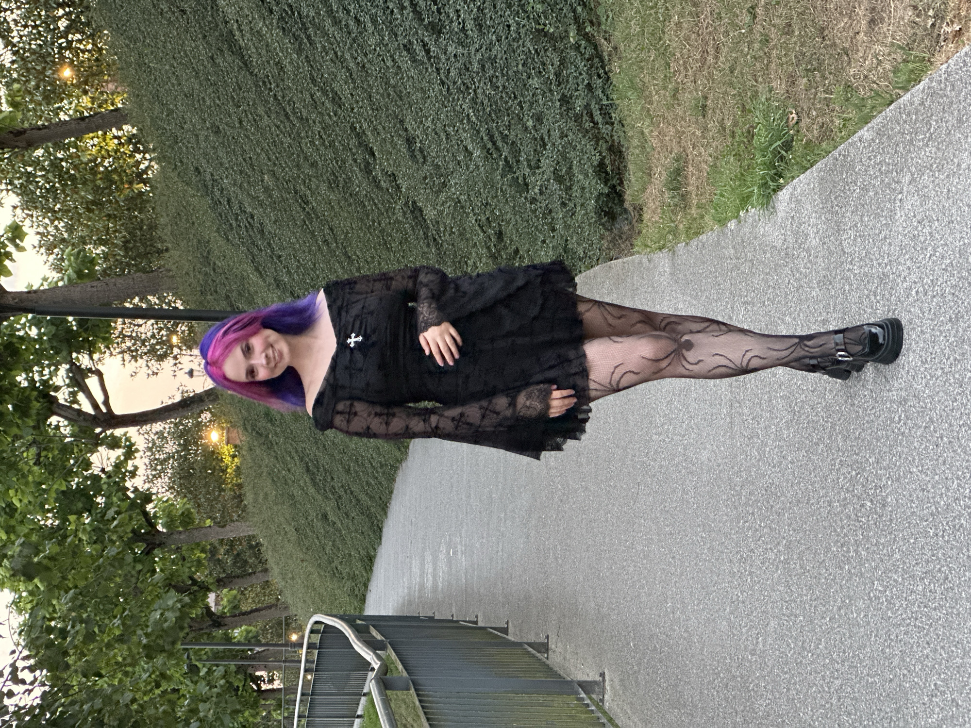 A young woman with purple and pink hair stands on a paved path next to a dense green hedge. She is wearing a black lace dress with long sleeves, sheer black patterned stockings, and black platform shoes. A small silver cross is pinned to her dress. Her right hand rests on her hip. The sun is setting, casting a warm glow through the trees on the left side of the image. The weather appears to be pleasant, with clear skies. The scene suggests an outdoor setting, possibly a park or a street in Oviedo, Spain, as indicated by the location context. The woman has a slight smile, conveying a sense of calm or confidence.