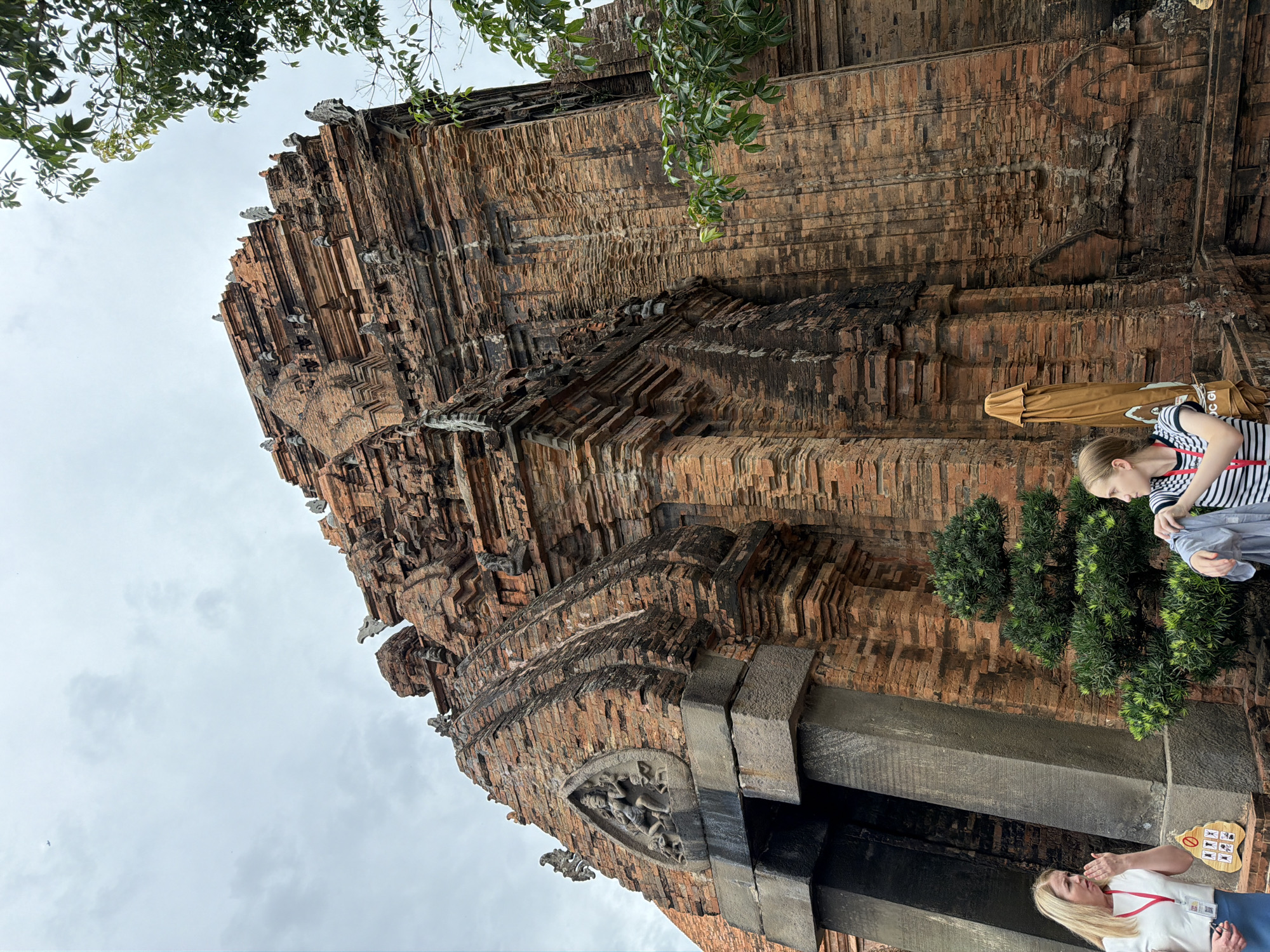 This image shows the Po Nagar Cham Towers in Nha Trang, Vietnam. The towers are made of red brick and are an ancient architectural style. The sky is overcast, and there are trees in the foreground. Two women are standing in front of the towers, and one of them is pointing. There is also a small, well-manicured tree.