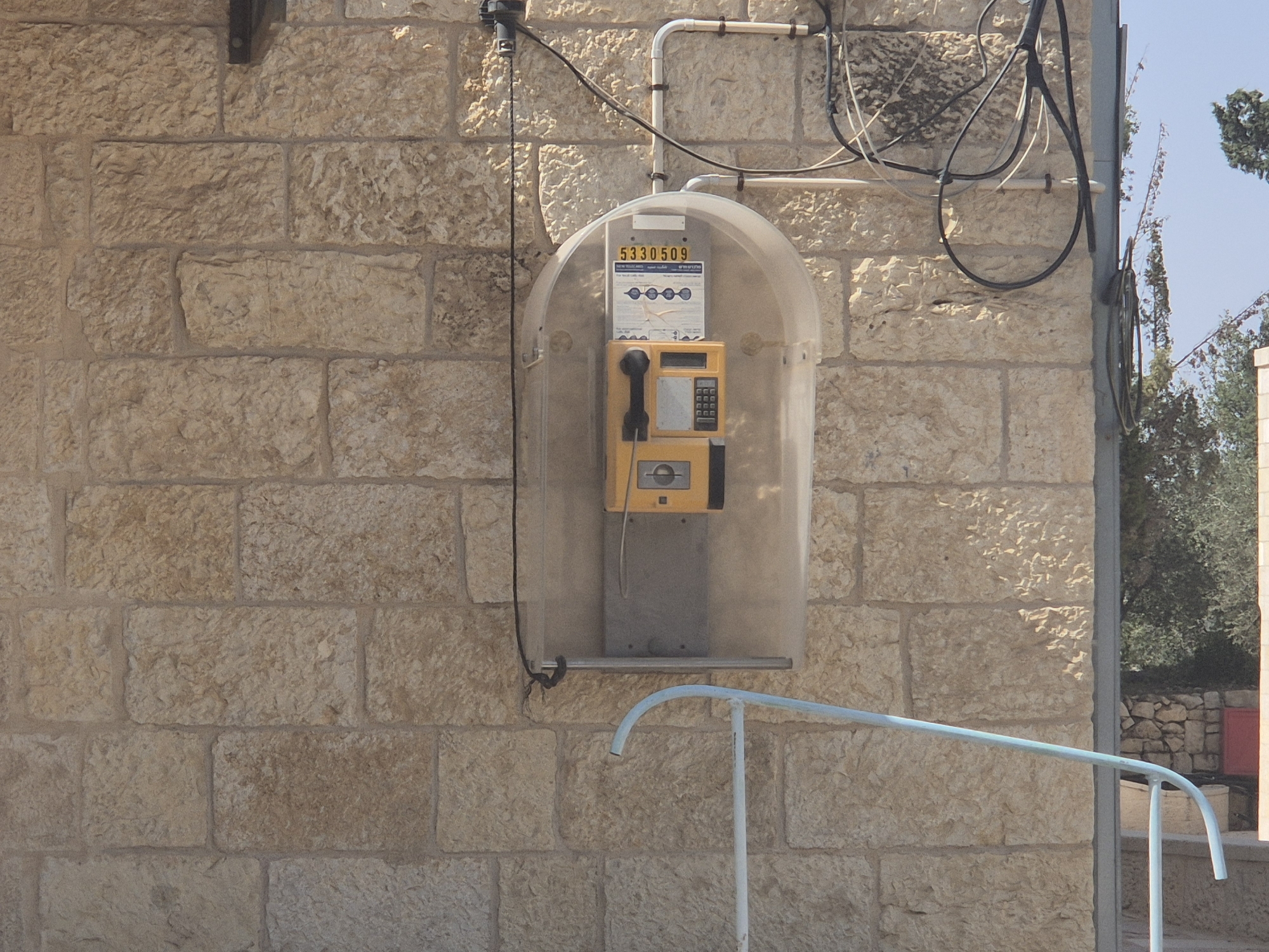 An exterior shot captures a weathered, light brown stone wall with visible mortar. Mounted on the wall is a clear plastic enclosure housing a yellow public payphone. The phone has a dark grey handset, a keypad, and a slot for coins or cards. Above the phone, a sign displays the number "5330509" and other text in Hebrew and English. Electrical wires, both black and white, are routed along the wall and into the enclosure. A light blue metal railing, curved at one end, extends from the bottom right of the frame upwards towards the left. In the background, trees and a blue sky are visible. The setting appears to be the historic village of Abu Gosh, Israel, a location known for its ancient architecture and cultural significance.