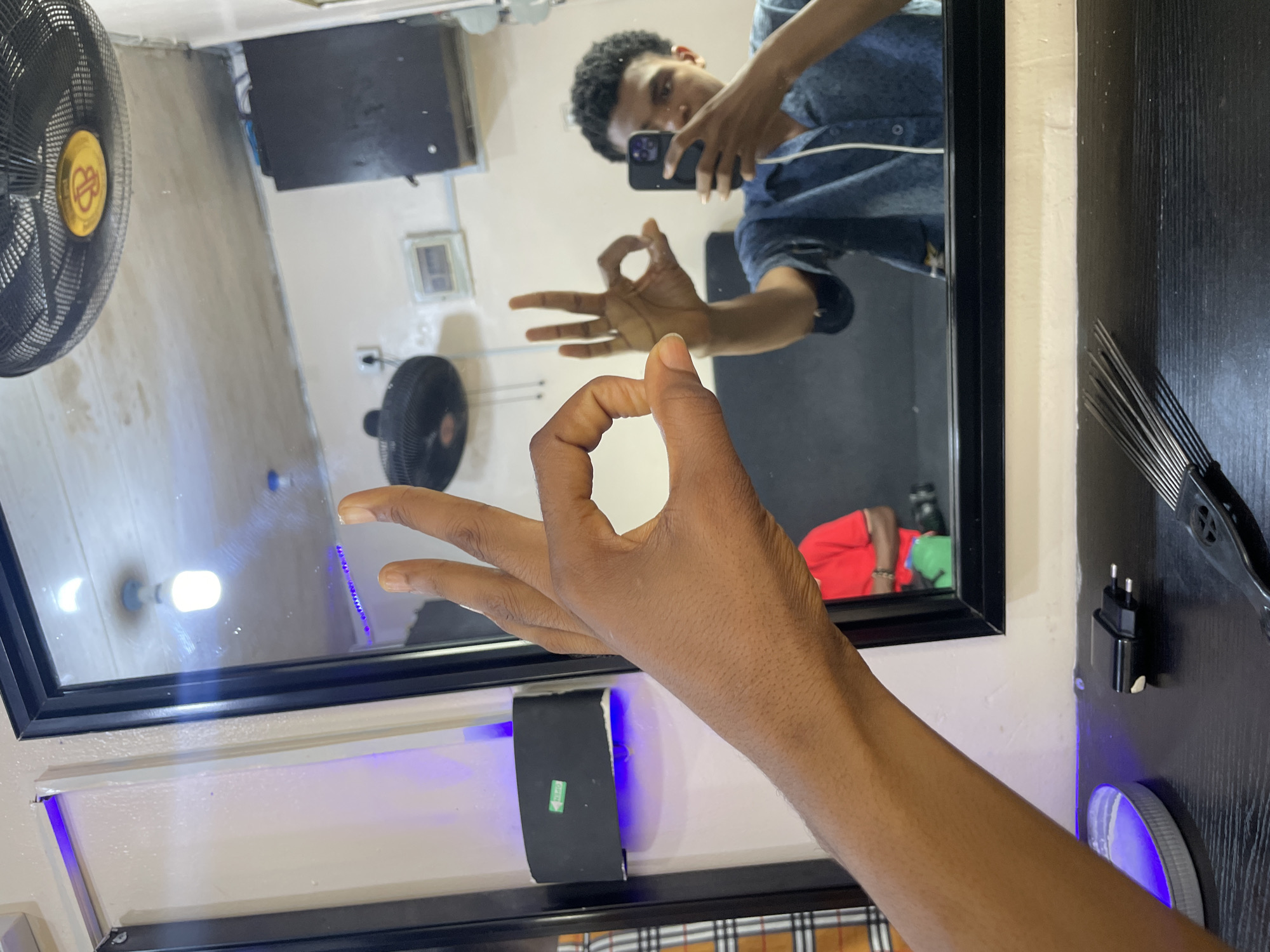 A person extends their right hand toward the camera, making an "OK" sign with the index finger and thumb. The arm is dark-skinned and the fingers are slender. The hand is in front of a black-framed mirror that reflects a person holding a smartphone taking a picture of themselves. The reflected person is wearing a blue button-down shirt. In the reflection, another person is visible wearing a red shirt and green pants. The background includes a black fan and a white wall. A hair pick and charger are hanging on a black wooden dresser to the right of the mirror. A light with a blue glow is positioned beneath the mirror. The location is Ado, Nigeria.